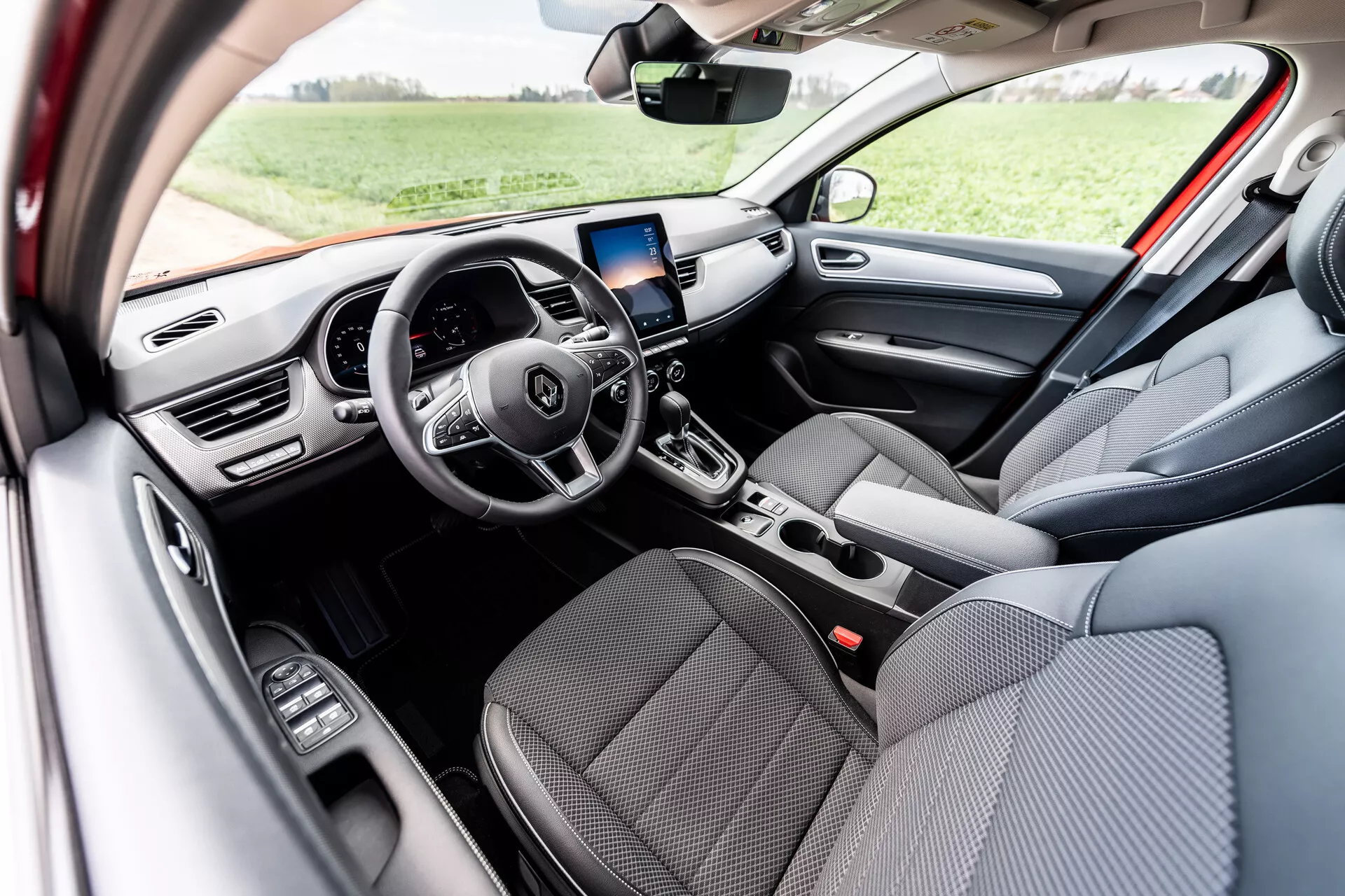 Vue intérieure du Renault Arkana 2021 mettant en avant des sièges avant en tissu gris, un volant multifonction à trois branches avec boutons de commande intégrés, affichage numérique du tableau de bord, écran d'infodivertissement avec navigation GPS au-dessus des bouches d'aération centrales, commandes manuelles de climatisation, levier de vitesse automatique, et commandes de lève-vitres électriques sur la porte côté conducteur, éclairée par la lumière naturelle du jour dans un cadre extérieur