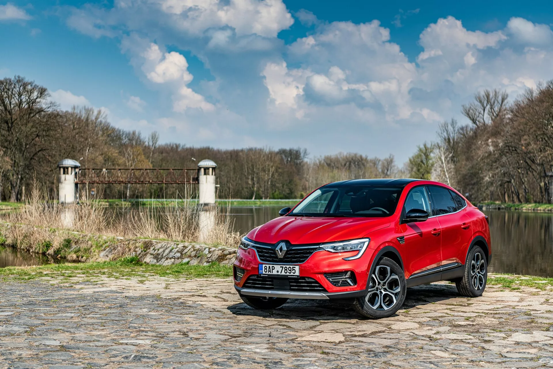 L'image montre une Renault Arkana 2021 garée sur un chemin pavé près d'une rivière ou d'un lac, avec de la végétation et des structures ressemblant à des tours de barrage ou de pont en arrière-plan. La voiture est capturée d'un angle bas, vue de trois quarts avant, mettant en avant la calandre avant, les phares à LED et une partie du profil latéral incluant la porte du conducteur et l'aile avant. Les caractéristiques de conception moderne, telles que la signature lumineuse nette des LED et les grandes roues en alliage, suggèrent un niveau de finition élevé. La peinture métallique réfléchissante ajoute à l'attrait esthétique, tandis que les possibles capteurs d'aide à la conduite et la technologie intégrée aux pare-chocs et aux rétroviseurs latéraux améliorent sa fonctionnalité.