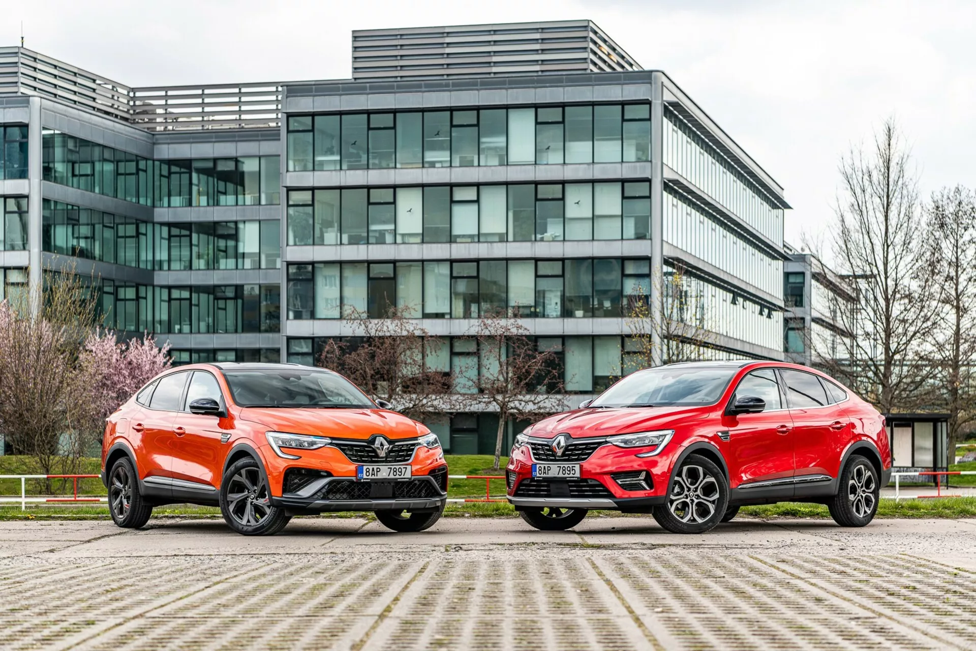 L'image montre deux véhicules de type SUV garés côte à côte devant un bâtiment de bureaux moderne à façade de verre. Le véhicule à gauche est peint en orange vif avec une finition métallique et présente une calandre avant distinctive, des phares LED nets, un capot sculpté et de grandes roues en alliage multi-brins avec des pneus à profil bas. La voiture à droite a une finition rouge métallique, avec une silhouette globale similaire, des phares nets, une calandre proéminente et de grandes roues en alliage stylées. Les deux véhicules présentent des lignes angulaires, des rétroviseurs aérodynamiques et des moulures inférieures contrastantes. La photo, prise à un angle bas près du niveau du sol, souligne l'environnement urbain et met en valeur le design, la présence et les caractéristiques technologiques avancées des véhicules.