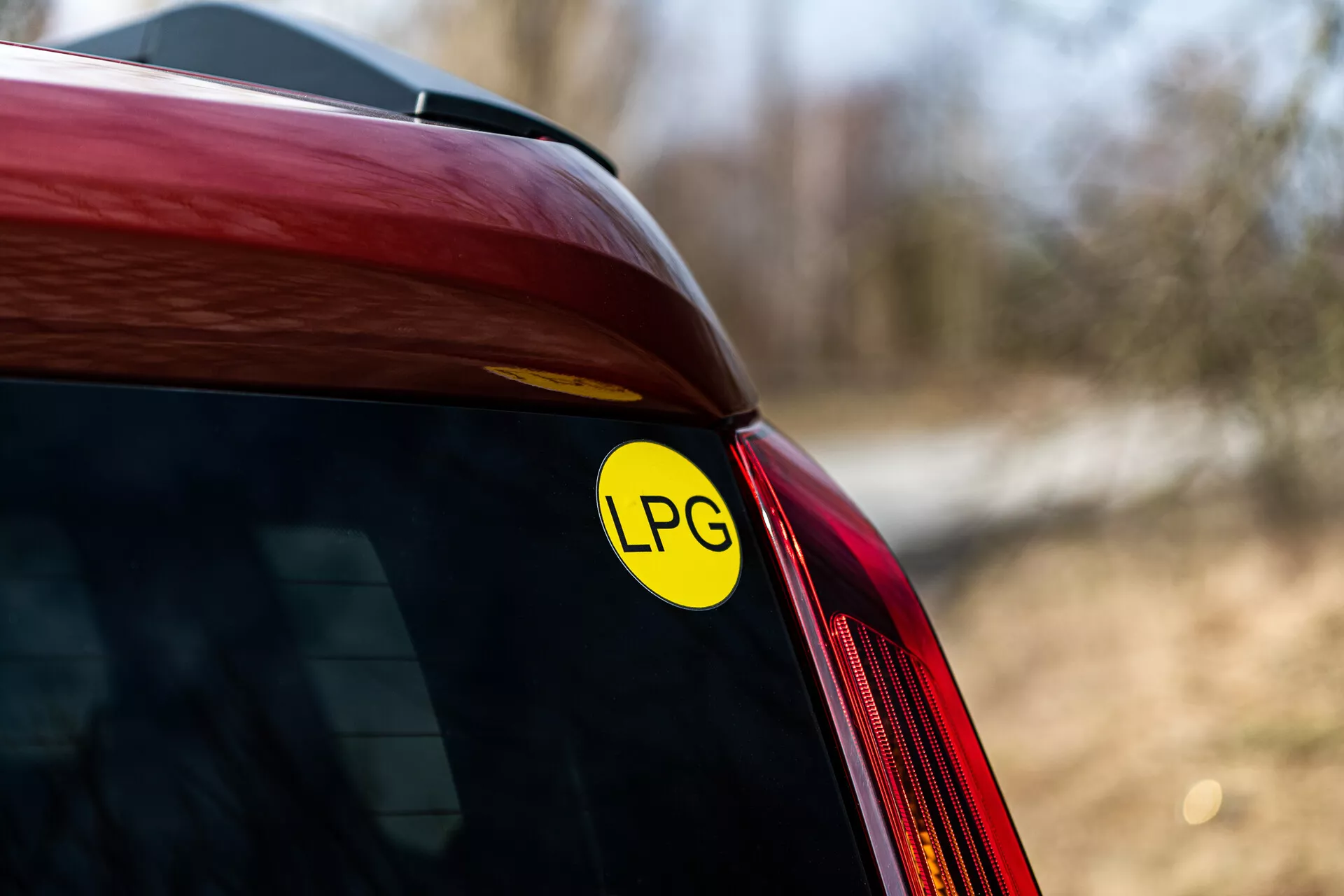 Gros plan sur l'angle arrière d'une Renault Arkana rouge de 2021 affichant l'autocollant GPL pour la capacité au gaz de pétrole liquéfié, centré sur la jonction du pare-brise arrière et du panneau latéral, sous la lumière naturelle en extérieur