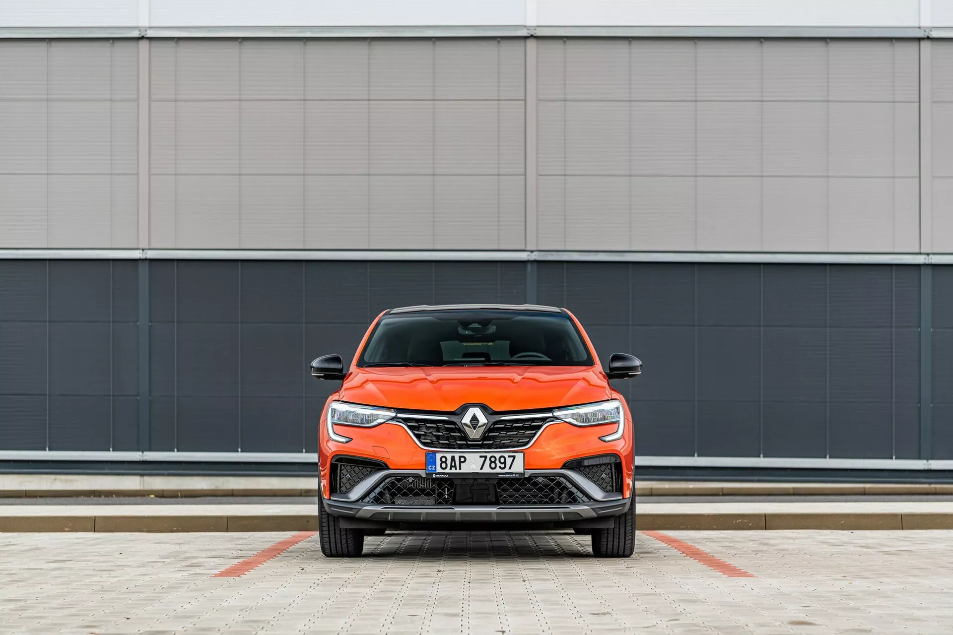 Vue frontale d'une voiture Renault orange vif, mettant en valeur son design moderne avec des phares élégants et une calandre avant, garée perpendiculairement à la caméra contre un fond de mur gris minimaliste dans une zone de stationnement avec des lignes.