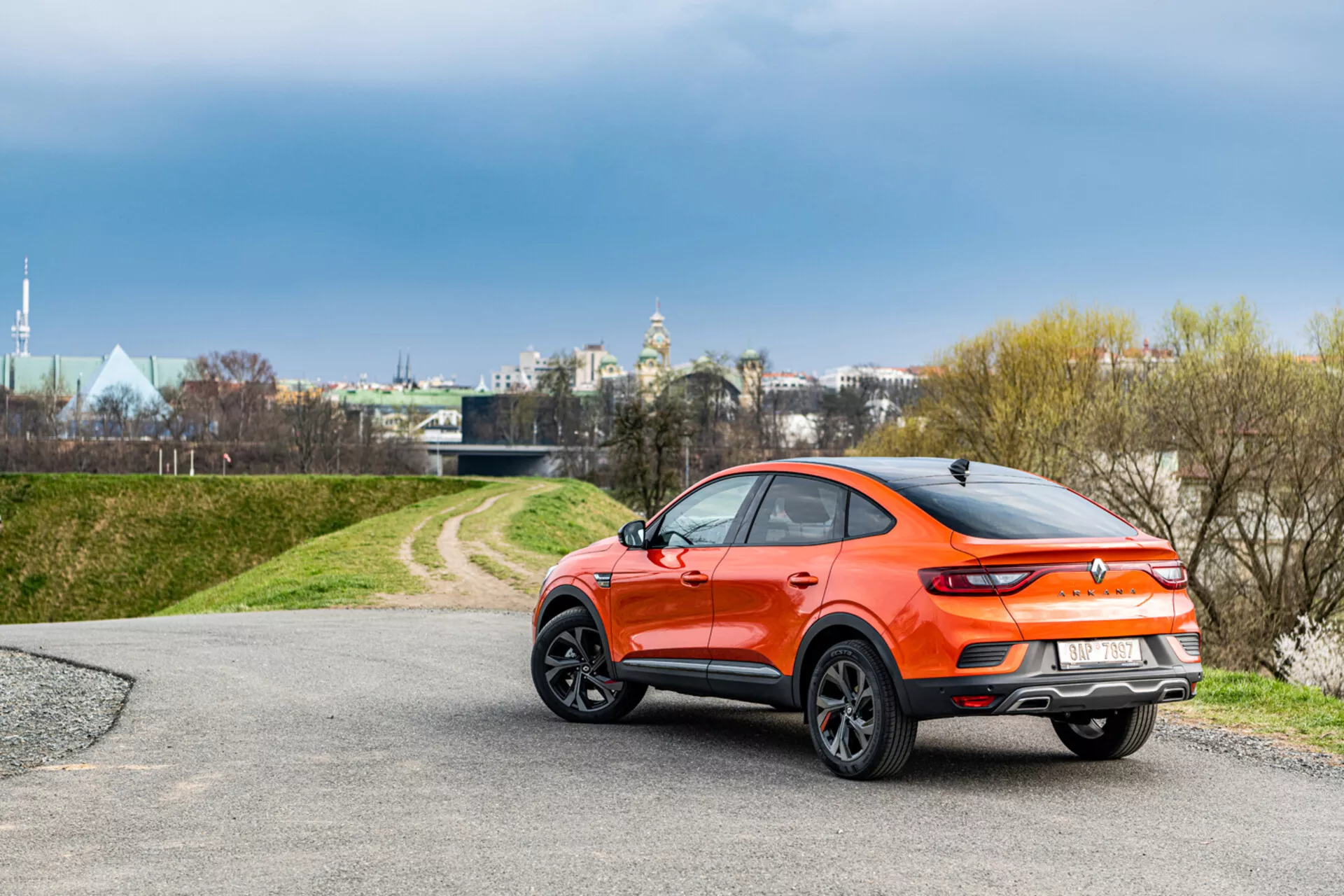 SUV crossover compact orange Renault Arkana de 2021 sous un angle droit montrant le profil et une vue partielle arrière, placé contre un décor naturel avec un éclairage doux qui met en valeur son design élégant de style coupé sportif et ses caractéristiques modernes.