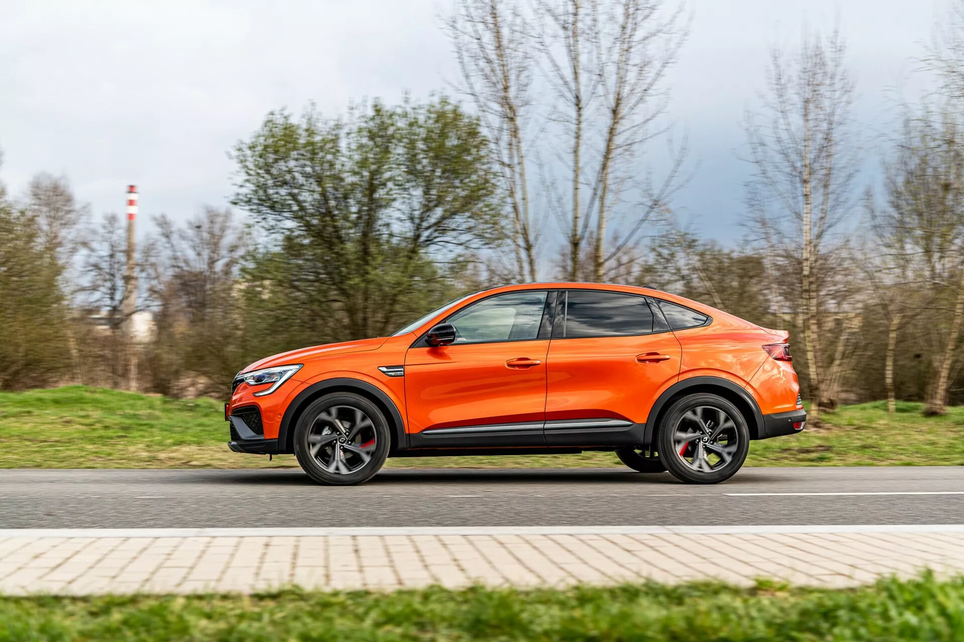 Photo dynamique de profil latéral d'un SUV crossover Renault Arkana 2021 orange en mouvement, capturée sous un angle bas mettant en valeur sa garde au sol et sa posture sportive. La longueur complète de la voiture est visible, mettant en évidence les lignes de design, les jantes en alliage, les portes latérales profilées et un toit de style coupé. Un léger flou de mouvement sur les roues et l'arrière-plan suggère une vitesse modérée, avec la voiture au point focal net. L'image met en avant le design moderne et aérodynamique du véhicule.