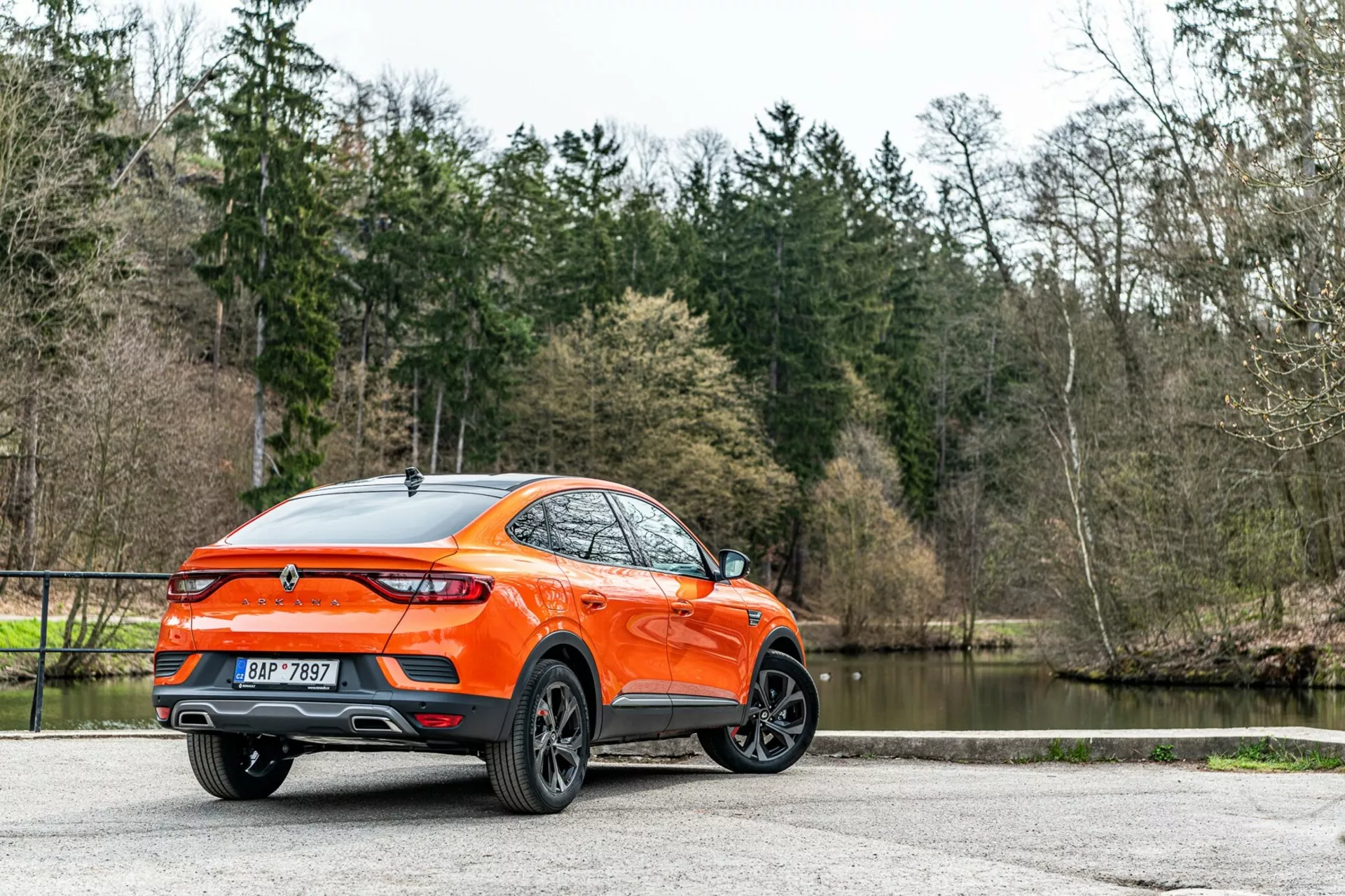 Renault Arkana 2021 vu sous un angle de trois quarts arrière, vue détaillée des profils latéral et arrière, présentant un crossover/SUV moderne avec plus de garde au sol et une ligne de toit de type coupé sport. La carrosserie orange vif se détache contre les arbres et le lac, mettant en valeur les roues en alliage au design multi-branche, les feux arrière à LED, le spoiler arrière, l'antenne aileron de requin, l'emblème central, les doubles sorties d'échappement, la finition de peinture brillante et les vitres arrière teintées. L'éclairage naturel diffus améliore la visibilité de l'extérieur bien entretenu, suggérant une attraction pour les consommateurs aux modes de vie actifs, garé sur une surface pavée.