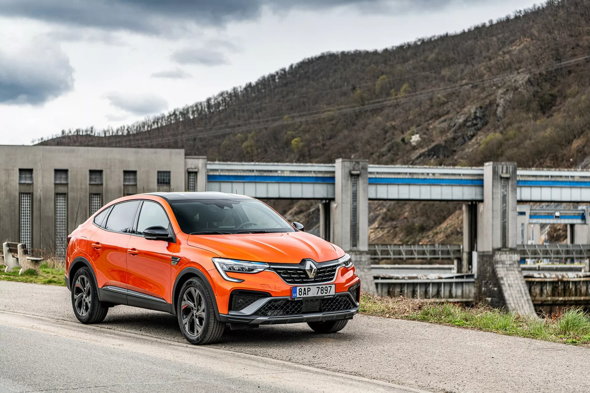 Un Renault Arkana 2021 crossover SUV moderne, de couleur orange vif, est garé le long d'une route, photographié sous un angle de profil avant mettant en valeur le côté conducteur. L'image montre la calandre avant de la voiture avec un grand emblème, des phares LED angulaires et élégants, des feux de jour, des entrées d'air et potentiellement des capteurs d'assistance à la conduite sur le pare-chocs inférieur, des arches de roue prononcées avec des roues en alliage à plusieurs branches et des rétroviseurs avec clignotants intégrés. Le véhicule, classé comme un SUV, a une posture surélevée avec une garde au sol accrue et une ligne de toit inclinée vers l'arrière typique des coupés. À l'arrière-plan, une infrastructure semblable à un barrage ou un système d'écluses peut être vue, situant la voiture dans un environnement industriel ou rural. La prise de vue est réalisée depuis un angle légèrement bas, soulignant la hauteur et la posture de la voiture.