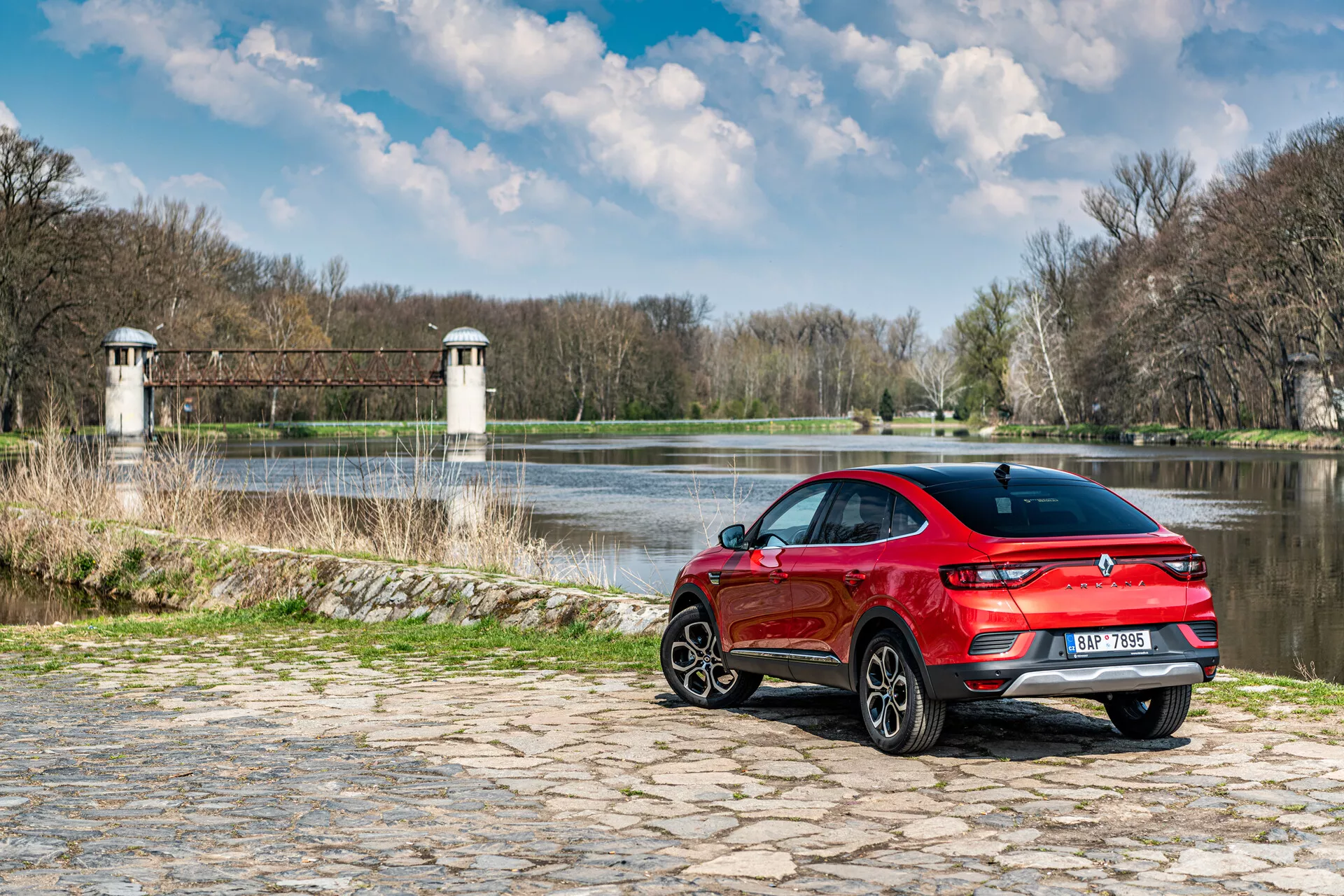 Renault Arkana 2021 garée sur pavés près de l'eau avec arrière-plan naturel, montrant vue arrière trois quarts, design sportif rouge et feux arrière LED