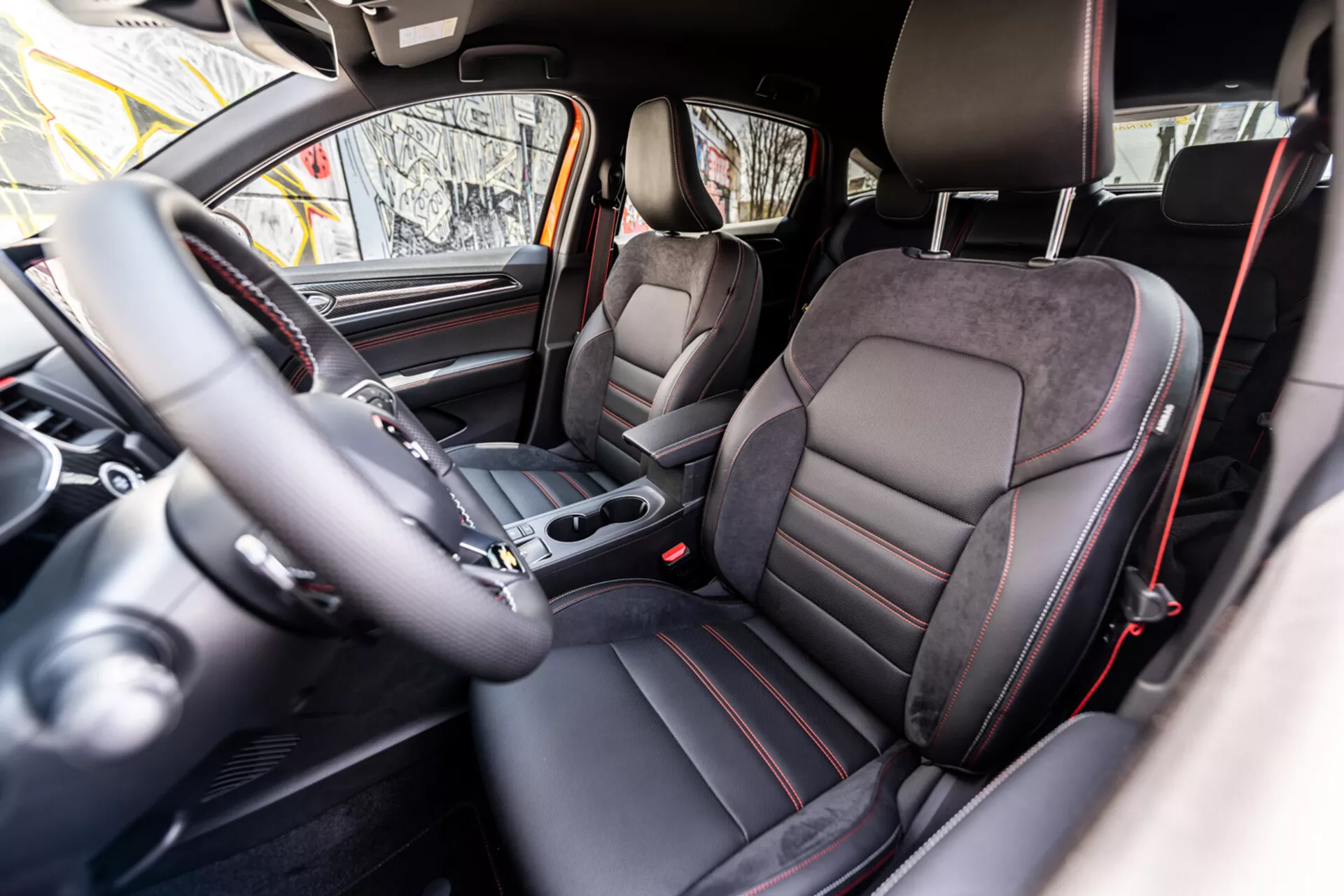 Intérieur de Renault Arkana 2021 montrant la cabine avant depuis le point de vue du siège arrière avec des sièges baquets de style sportif en cuir et Alcantara, volant à conduite à gauche avec commandes intégrées, levier de changement de vitesse automatique sur la console centrale, tableau de bord analogique, écran intégré du système d'infodivertissement et schéma de couleurs sombres premium avec accents métalliques