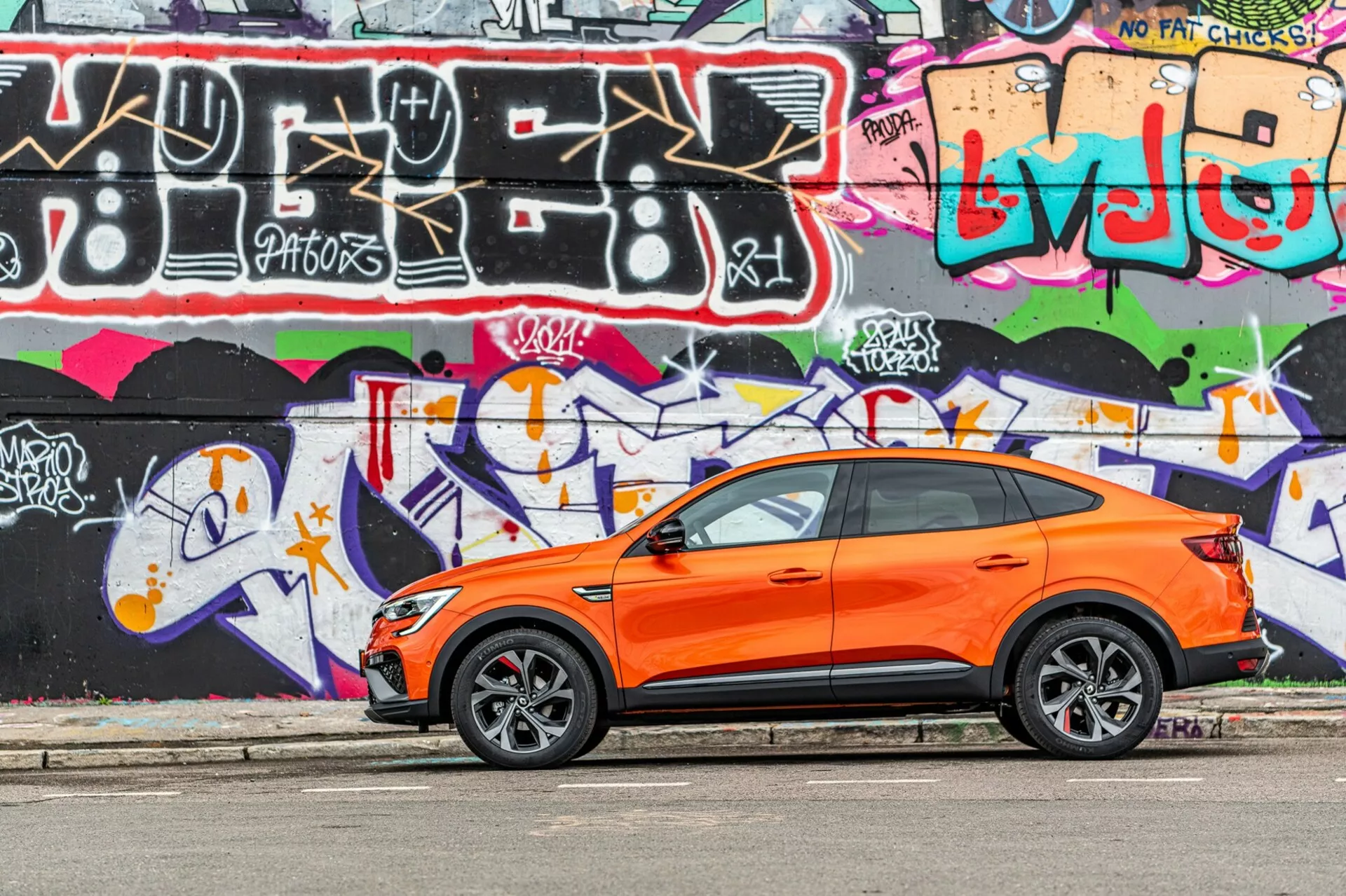 L'image présente un crossover SUV Renault Arkana 2021 vibrant orange, garé parallèlement à une rue avec un mur de graffitis coloré en arrière-plan. Capturée sous un angle de vue latérale, la photographie met en valeur le design sportif et moderne du véhicule avec les portes avant et arrière du côté conducteur visibles, les fenêtres latérales, les rétroviseurs latéraux et une ligne de toit inclinée. Les roues en alliage stylisées avec des pneus à profil bas suggèrent une focalisation sur les performances routières. Les poignées d'entrée sans clé sur les portes avant laissent présager un système d'entrée et de démarrage sans clé. L'image est prise en plein jour, soulignant le contraste du corps orange contre les graffitis, montrant une carrosserie bien entretenue et nette dans un environnement urbain et jeune.