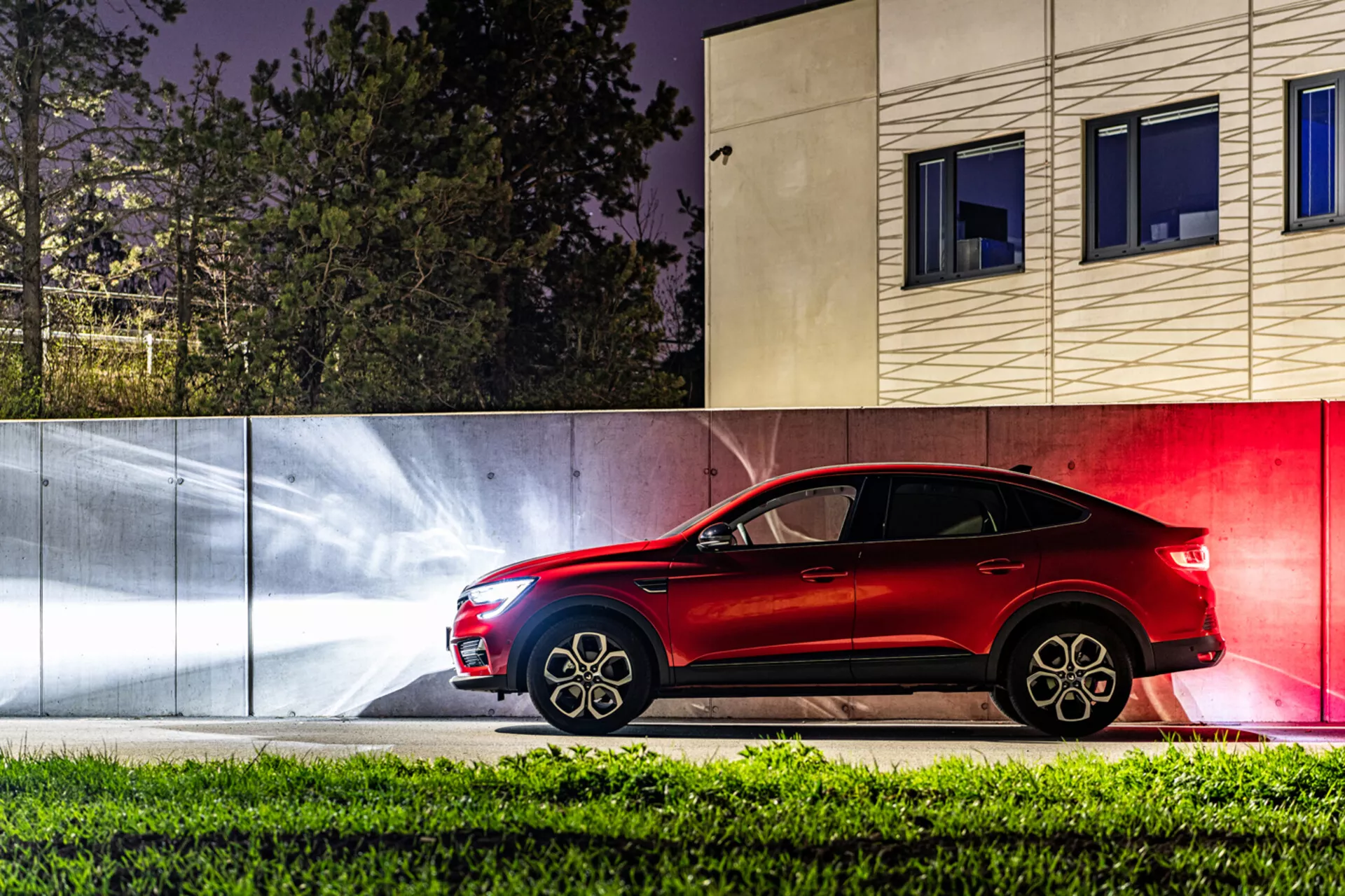 Vue de profil du Renault Arkana 2021 de nuit, mettant en lumière sa couleur rouge élégante, son design sportif et ses fonctionnalités avancées sur un fond urbain stylisé