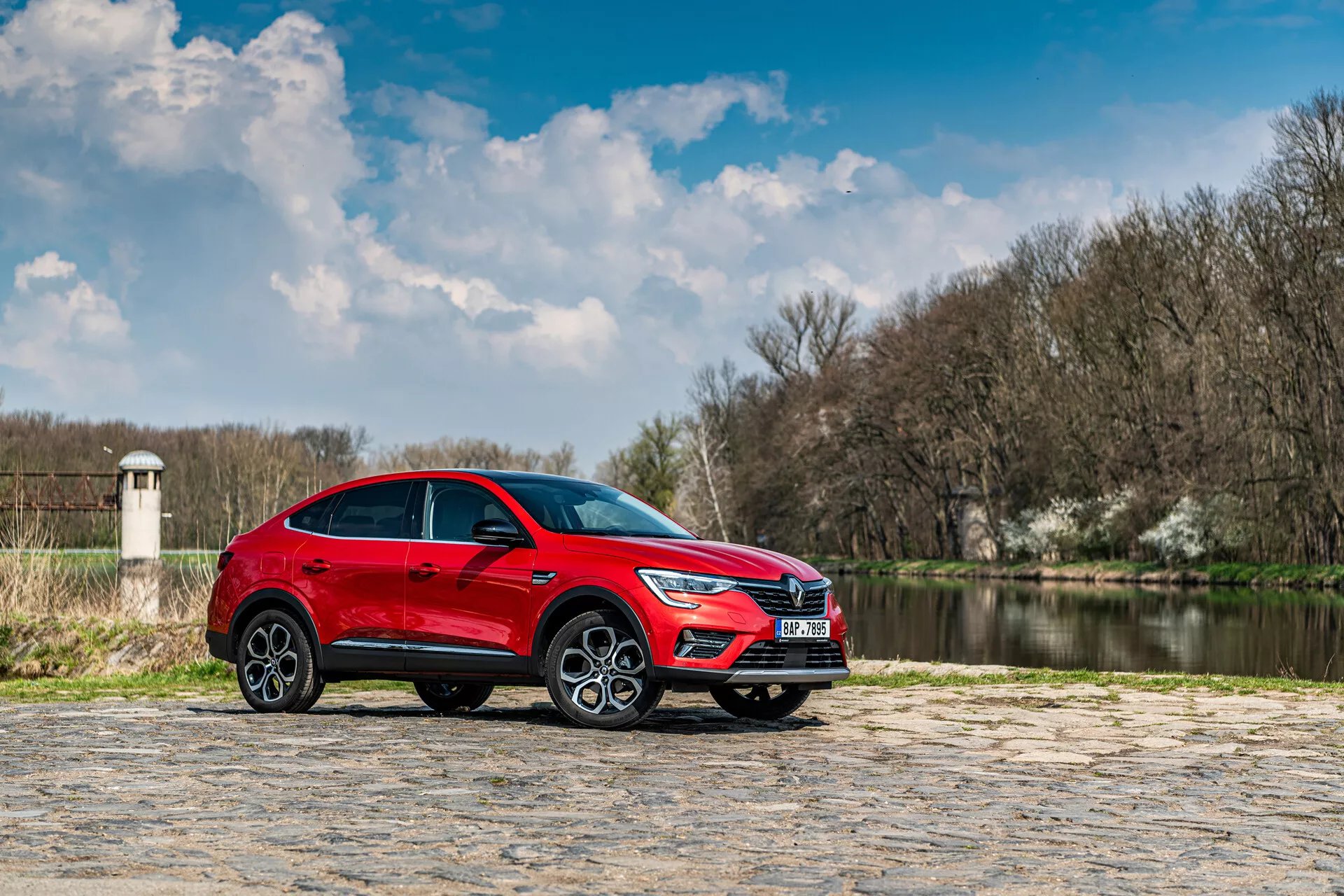 SUV rouge Renault Arkana 2021 garé sur un pavé en pierre avec de l'eau et des arbres à l'arrière-plan sous un ciel partiellement nuageux, vue de profil mettant en avant son design élégant et ses caractéristiques dynamiques