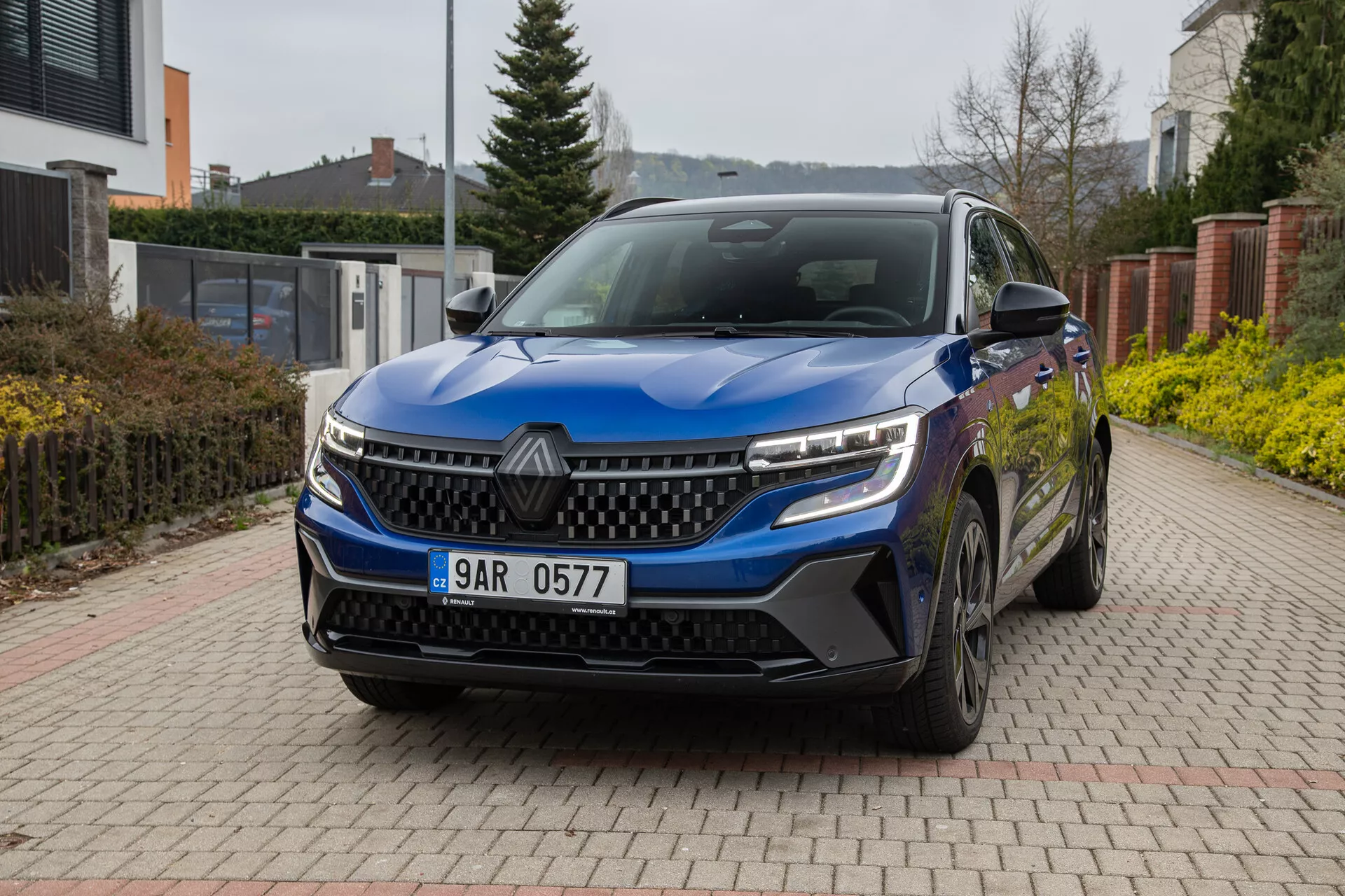Renault Austral 2022 en bleu, présentant son angle avant-trois-quarts sur une allée pavée de briques avec un cadre résidentiel en arrière-plan, comportant des phares LED, une grille distinctive, des capteurs avant, des roues en alliage et des rétroviseurs latéraux avec clignotants intégrés sous une lumière naturelle nuageuse.