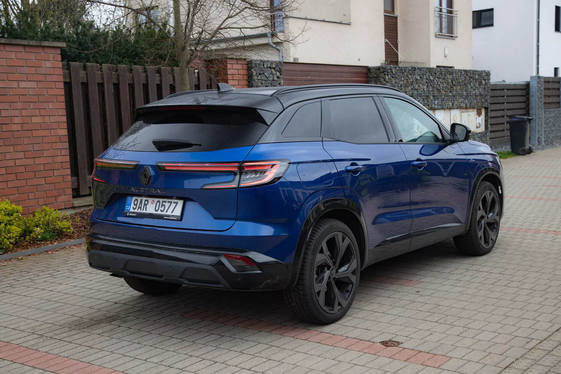 SUV moderne Renault Austral 2022 en bleu métallique brillant garé sur une allée pavée à côté d'un bâtiment résidentiel, montrant son aileron arrière sportif, ses feux arrière LED, ses roues en alliage de couleur sombre, ses vitres arrière teintées et ses capteurs de stationnement depuis un angle arrière de trois quarts