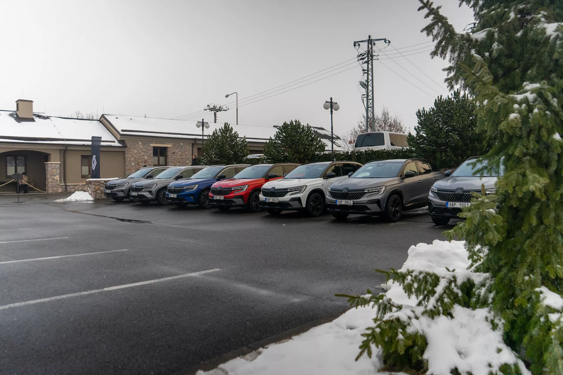 Alignement de voitures stationnées, mélange de berlines et de SUVs sous une légère chute de neige, indiquant des conditions météorologiques froides, avec des profils latéraux visibles incluant portes, rétroviseurs et roues, pris depuis un angle de trottoir surélevé