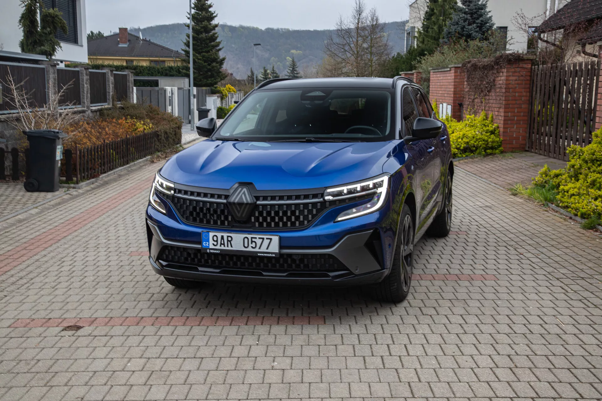 Vue de face en angle d'un SUV Renault Austral 2022 garé dans une allée résidentielle, montrant la grille, les phares LED, les feux de jour, le pare-chocs avec prises d'air, les capteurs de stationnement, l'emblème, les roues en alliage, les rétroviseurs latéraux avec clignotants et la plaque d'immatriculation.