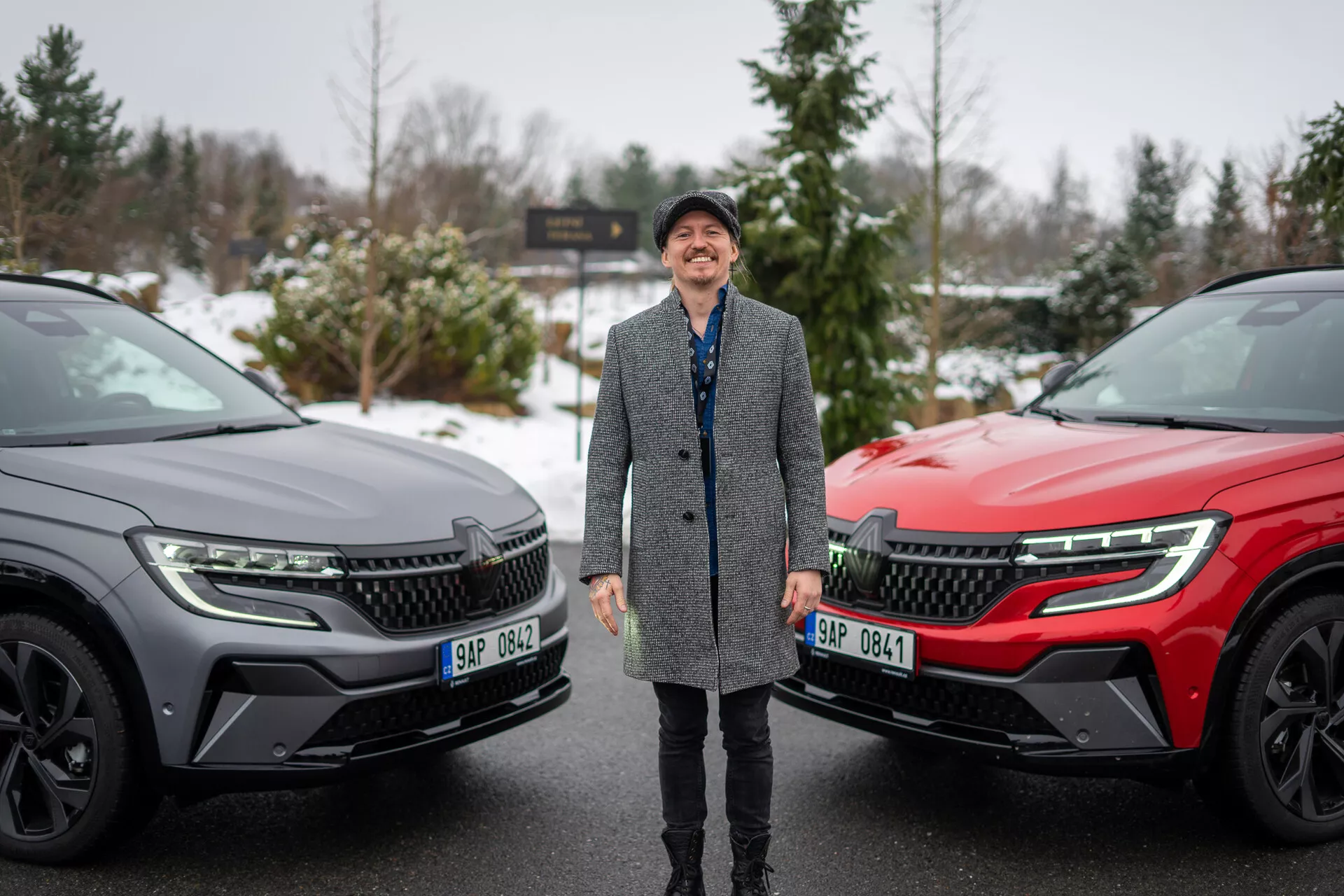 Personne souriante entourée de deux SUV Renault Austral 2022 dans un cadre hivernal, mettant l'accent sur le design moderne des véhicules et les caractéristiques technologiques avancées, avec un focus sur la grille avant, les phares LED et le profil latéral élégant.