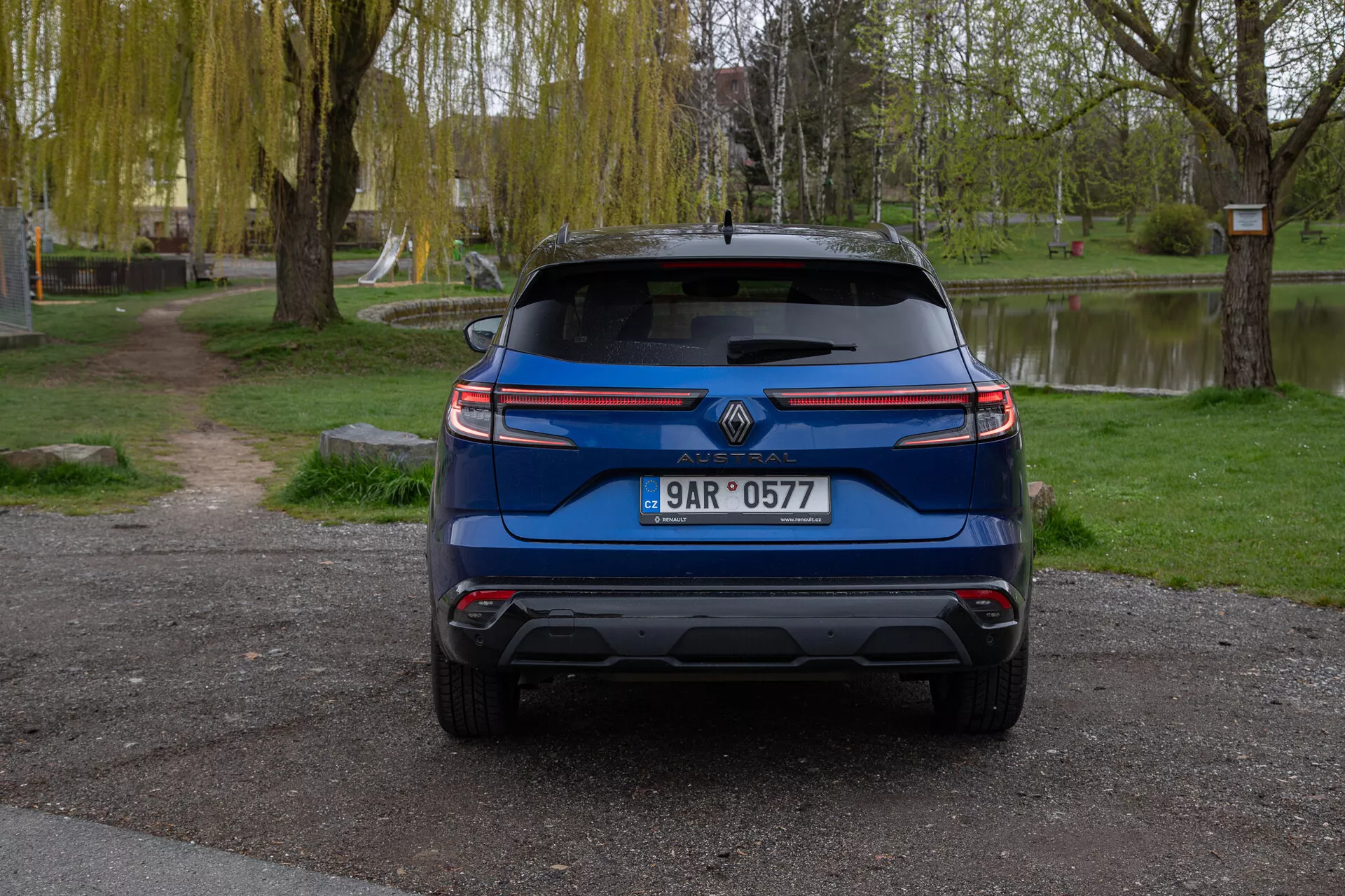 Vue arrière d'une Renault Austral 2022 bleue, garée à l'extérieur montrant des caractéristiques comprenant le logo de la marque, plaque d'immatriculation, feux arrière modernes, pare-brise arrière avec essuie-glace, pare-chocs arrière conçu avec des capteurs potentiels d'aide au stationnement, embouts d'échappement visibles, réflecteurs ou capteurs, et un spoiler arrière pour améliorer l'aérodynamique, le tout sur un fond serein semblable à un parc avec des arbres, de l'herbe et un plan d'eau.