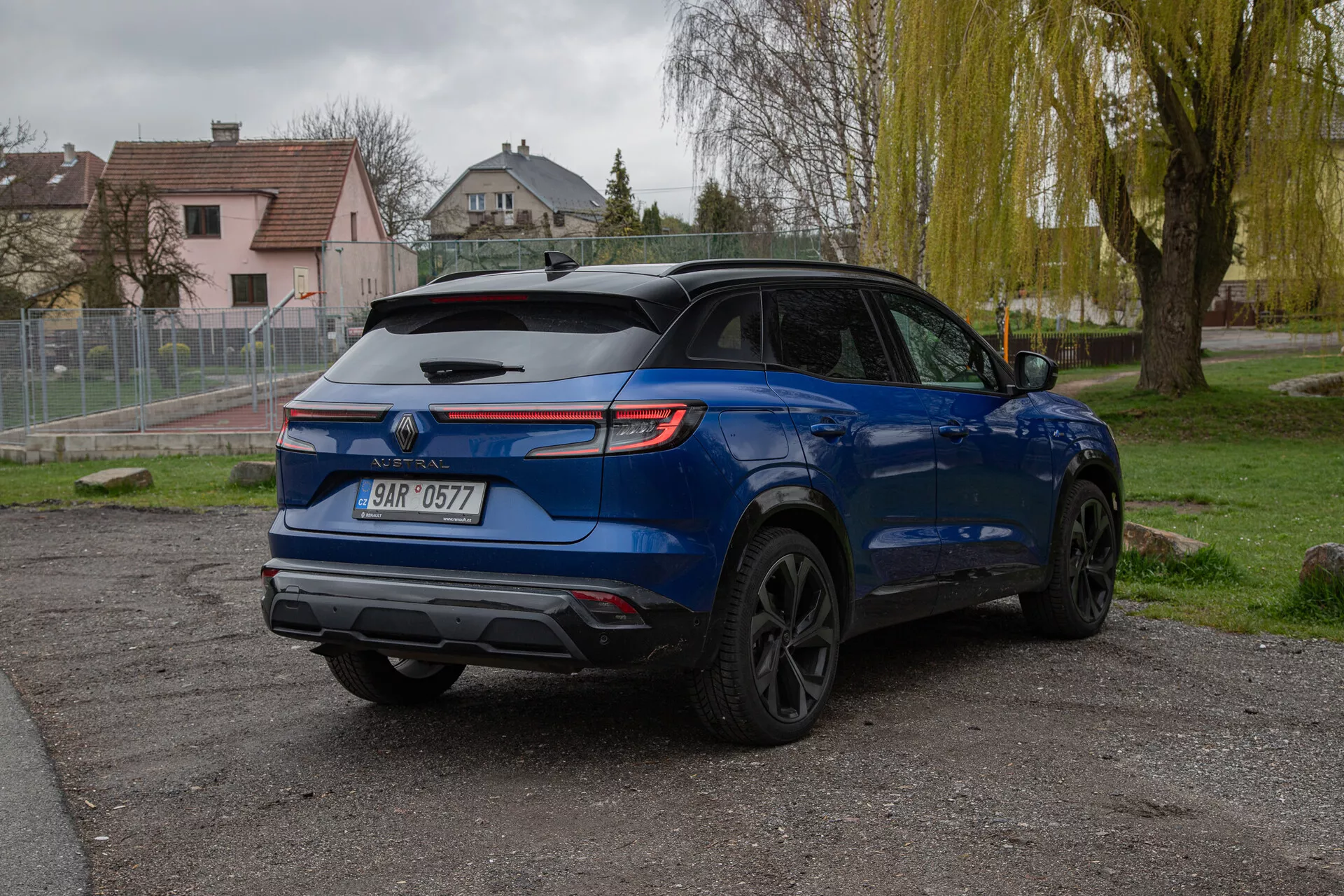 Image d'un crossover/SUV Renault Austral 2022 bleu garé à l'extérieur sur une surface non pavée avec un arrière-plan de gazon et d'arbres dans un cadre suburbain ou semi-rural, sous un éclairage doux de jour nuageux montrant une vue arrière de trois quarts mettant en avant son hayon avec pare-brise arrière et essuie-glace, spoiler, plaque d'immatriculation montée centralement sous l'emblème, feux arrière LED, doubles tuyaux d'échappement, porte-bagages, jantes en alliage noir, design de carrosserie sculpté et caméra de recul potentielle.