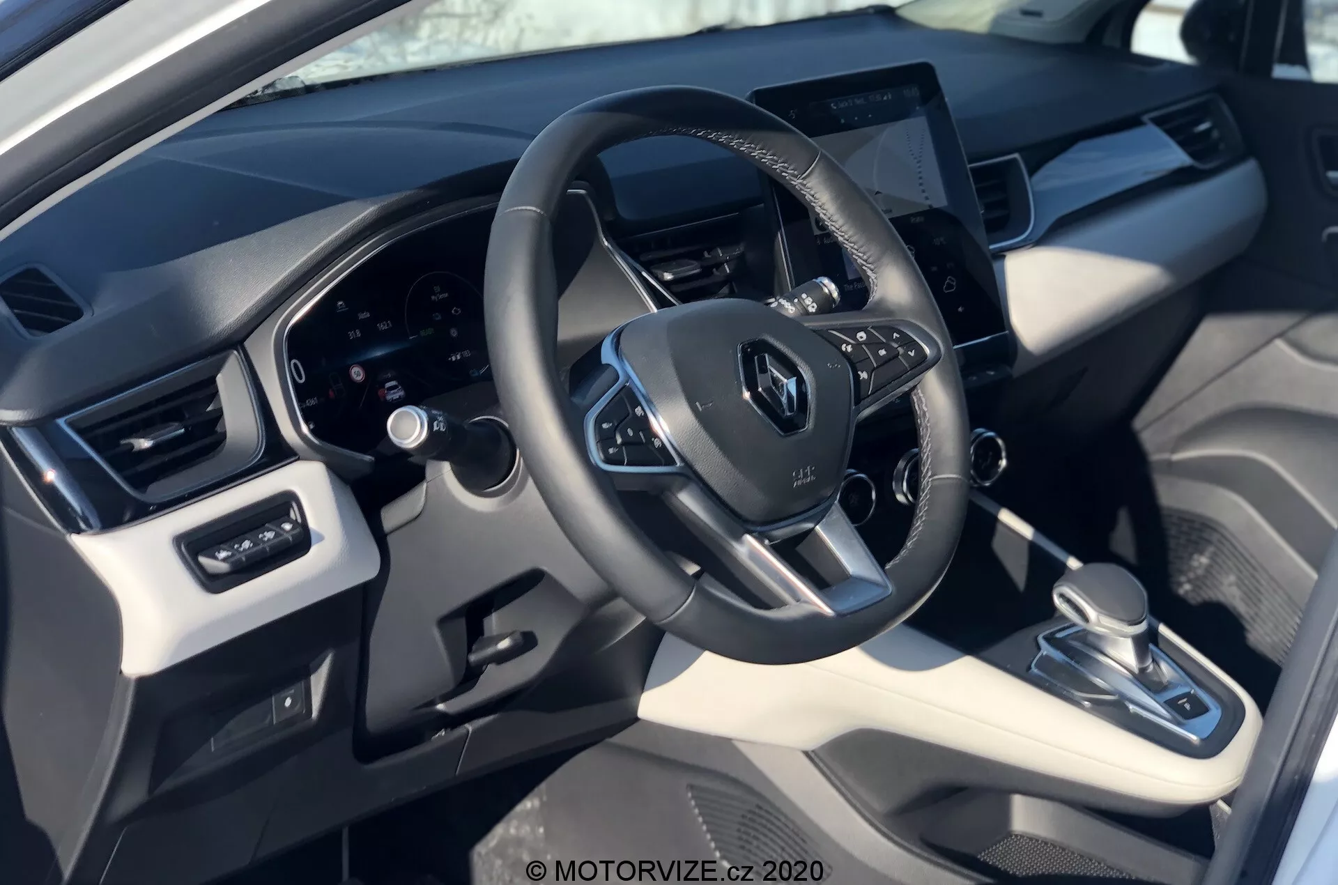 Vue intérieure de la Renault Captur 2020 depuis la perspective du conducteur, montrant le volant avec le logo Renault, les commandes intégrées, le tableau de bord numérique et un système d'infodivertissement à écran tactile centralement situé. L’image met en évidence un mélange de matériaux en cuir synthétique et en plastique avec des accents métalliques, une transmission automatique, un système de contrôle climatique et des réglages des vitres et des rétroviseurs électriques.