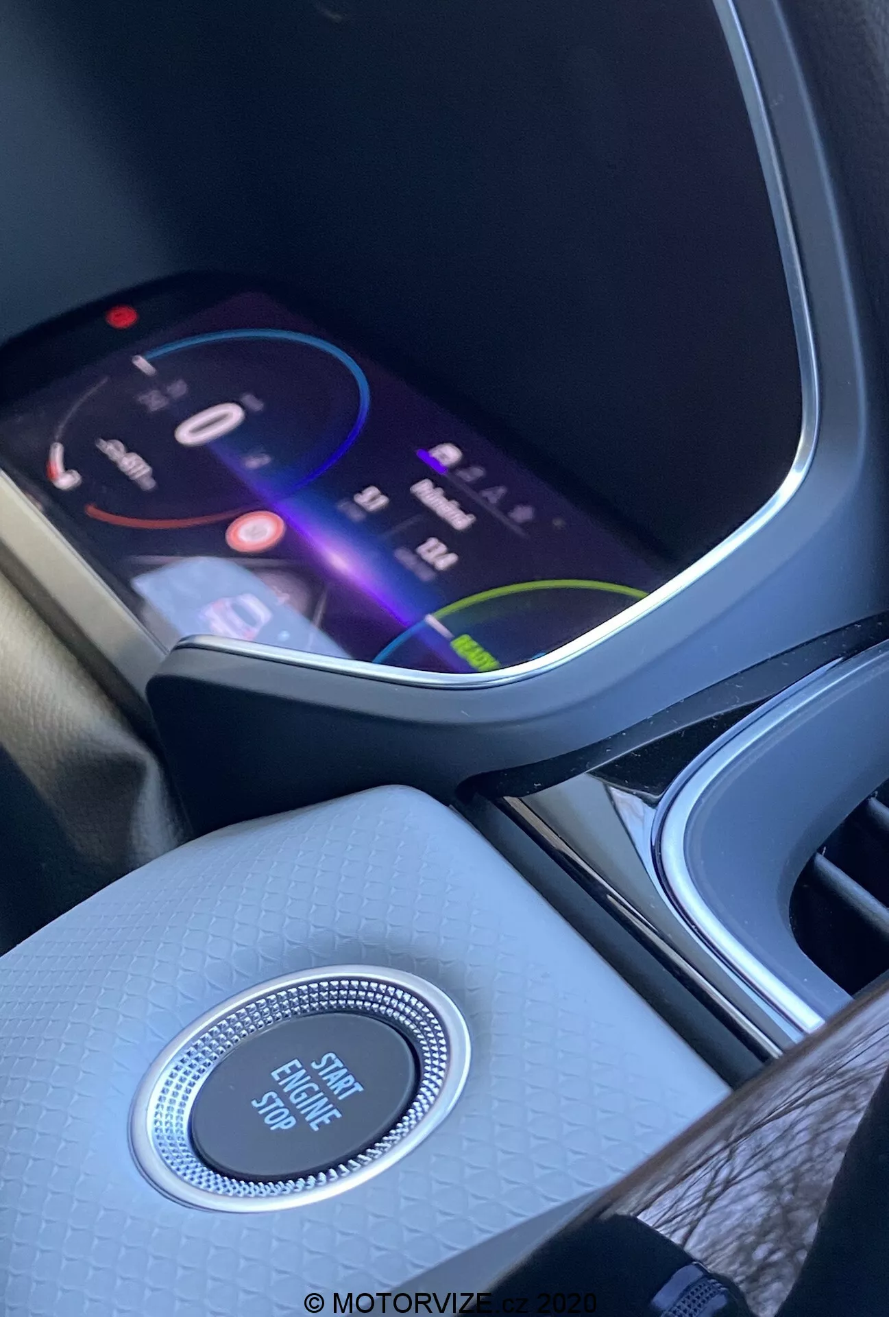 Intérieur de la Renault Captur 2020 mettant en avant la console centrale et le tableau de bord depuis la perspective du siège avant. L'écran d'infodivertissement vif affiche une interface colorée suggérant des fonctionnalités multimédia avancées, y compris la navigation, la lecture de médias et les options de connectivité. En dessous de l'écran, un bouton de commande circulaire à finition chromée marqué 'Engine Start Stop' met en évidence le système de démarrage sans clé. La console centrale présente une finition texturée de haute qualité, améliorant l'ambiance intérieure. À gauche, un porte-gobelet ou un compartiment de rangement partiellement visible.
