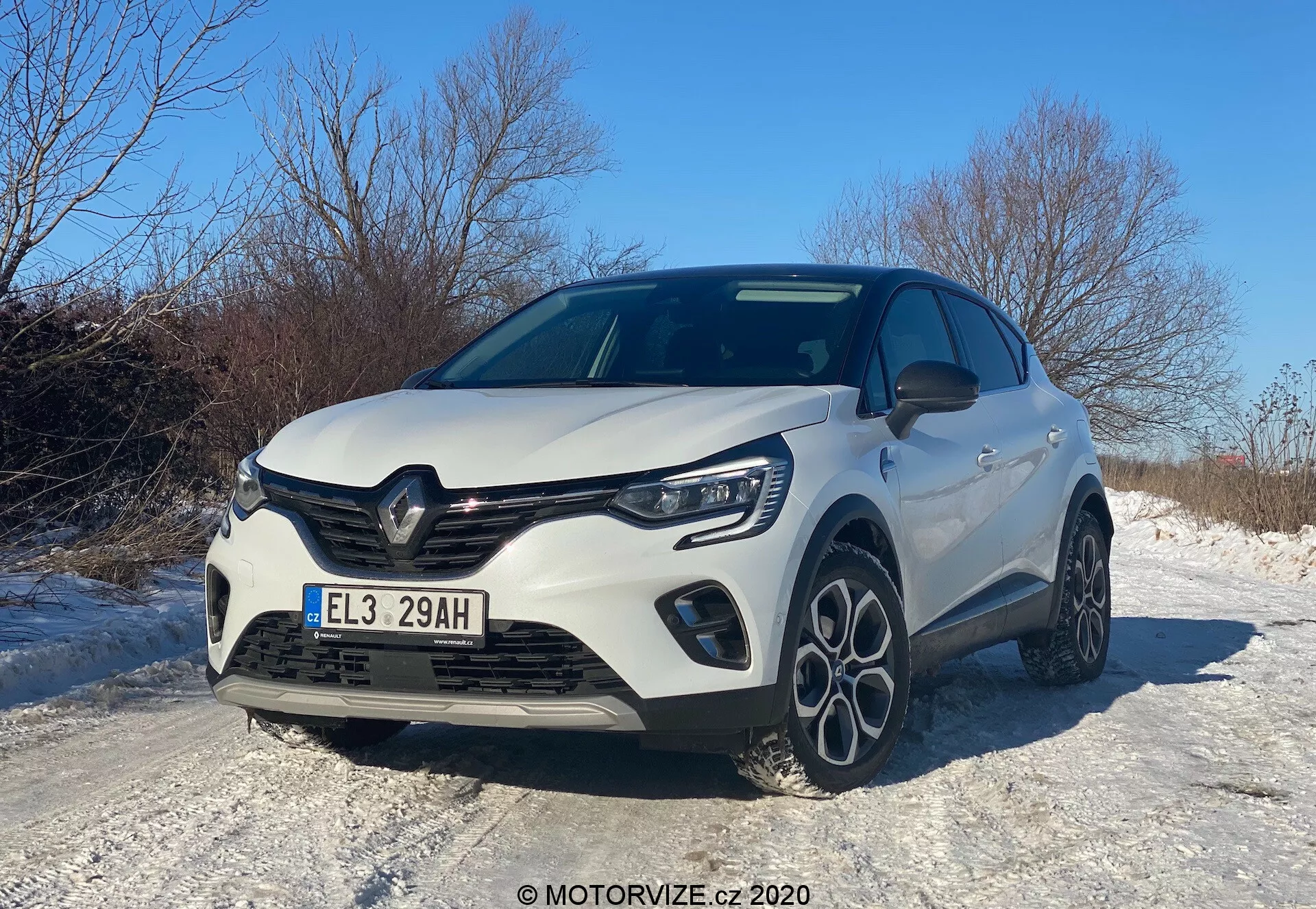 Véhicule hatchback moderne Renault Captur 2020 capturé sous un angle de trois quarts avant bas, montrant la calandre avant, le capot, les phares à LED, le pare-chocs avant avec des phares antibrouillard intégrés et un profil latéral partiel. Les éléments de design incluent une calandre proéminente avec l'emblème du constructeur, des lignes sculptées sur le capot et des jantes en alliage à plusieurs branches, le tout sous un ciel clair de jour, reflétant une esthétique sportive et aérodynamique, mettant peut-être en évidence une version améliorée ou un pack sport. La voiture est située sur une route enneigée, suggérant des caractéristiques appropriées pour la conduite hivernale.