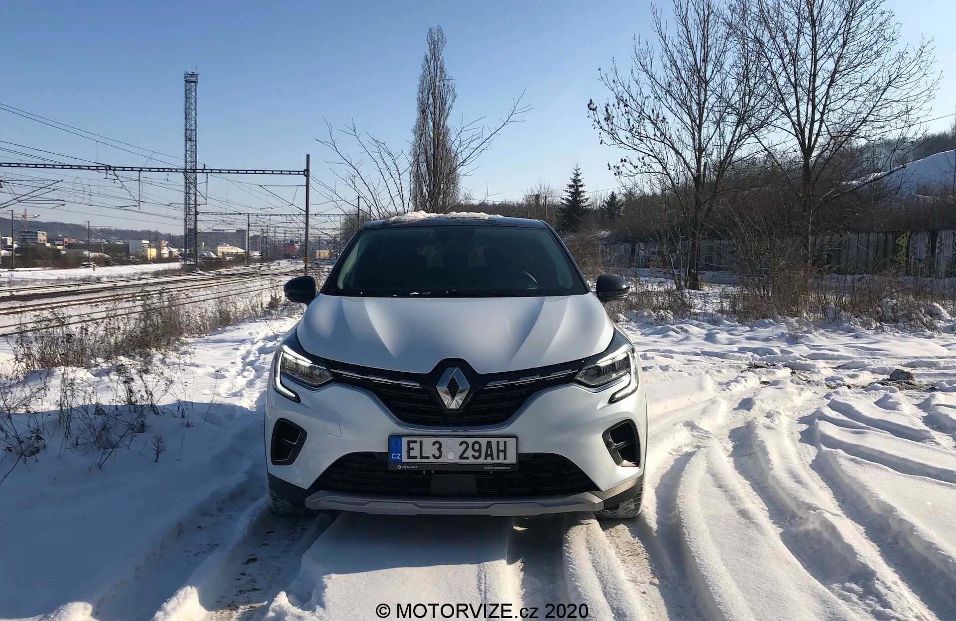 Vue frontale directe du Renault Captur 2020 sur une route couverte de neige, montrant en détail la calandre avant, les phares, le capot sculpté et le pare-chocs avant en pleine journée. Les traces de pneus sont visibles, témoignant d'un mouvement récent. Le design du véhicule suggère une aérodynamique stylée, avec un emblème de marque proéminent et un système d'éclairage avancé. Des capteurs ou caméras d'assistance à la conduite pourraient être intégrés dans le pare-chocs avant.