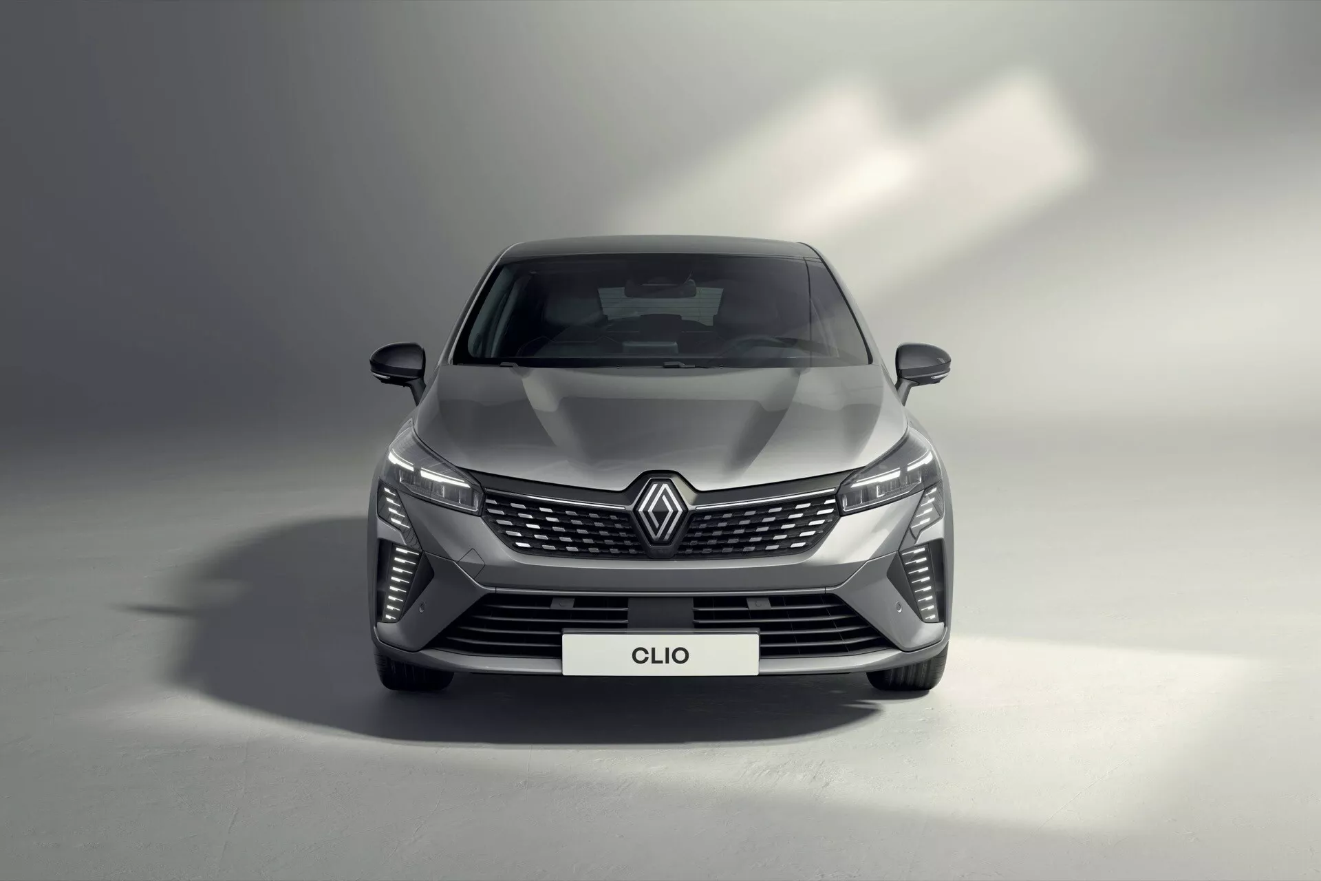 Vue frontale de la Renault Clio V-facelift 2023 montrant des phares LED, une grille large à motif diamant avec emblème de la marque, un pare-chocs au design aérodynamique avec entrées d'air et une finition métallique de haute qualité dans un cadre de studio