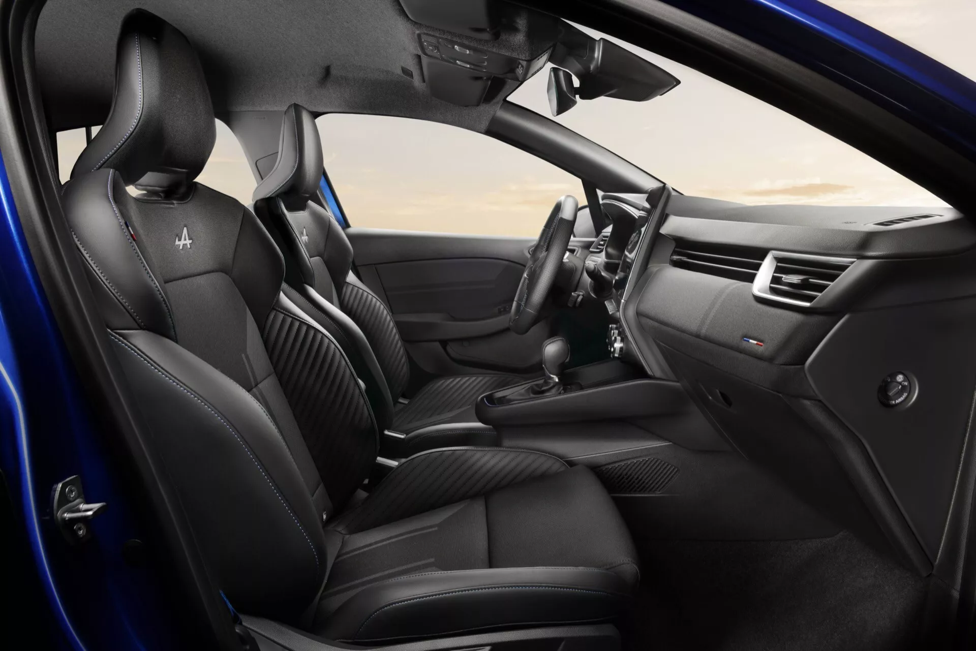 Intérieur du Renault Clio 2023 (V-Facelift) avec sièges avant en cuir noir, volant multifonction et système d'infodivertissement central, vu sous un angle élevé montrant la porte côté conducteur ouverte et les détails du tableau de bord