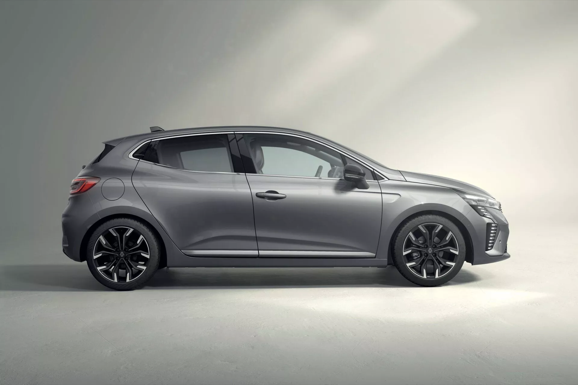 Vue de profil latéral de la moderne hatchback Renault Clio V-Facelift 2023 mettant en lumière la posture basse, le design des roues, la couleur gris métallisé et l'aérodynamisme élégant dans un cadre de studio