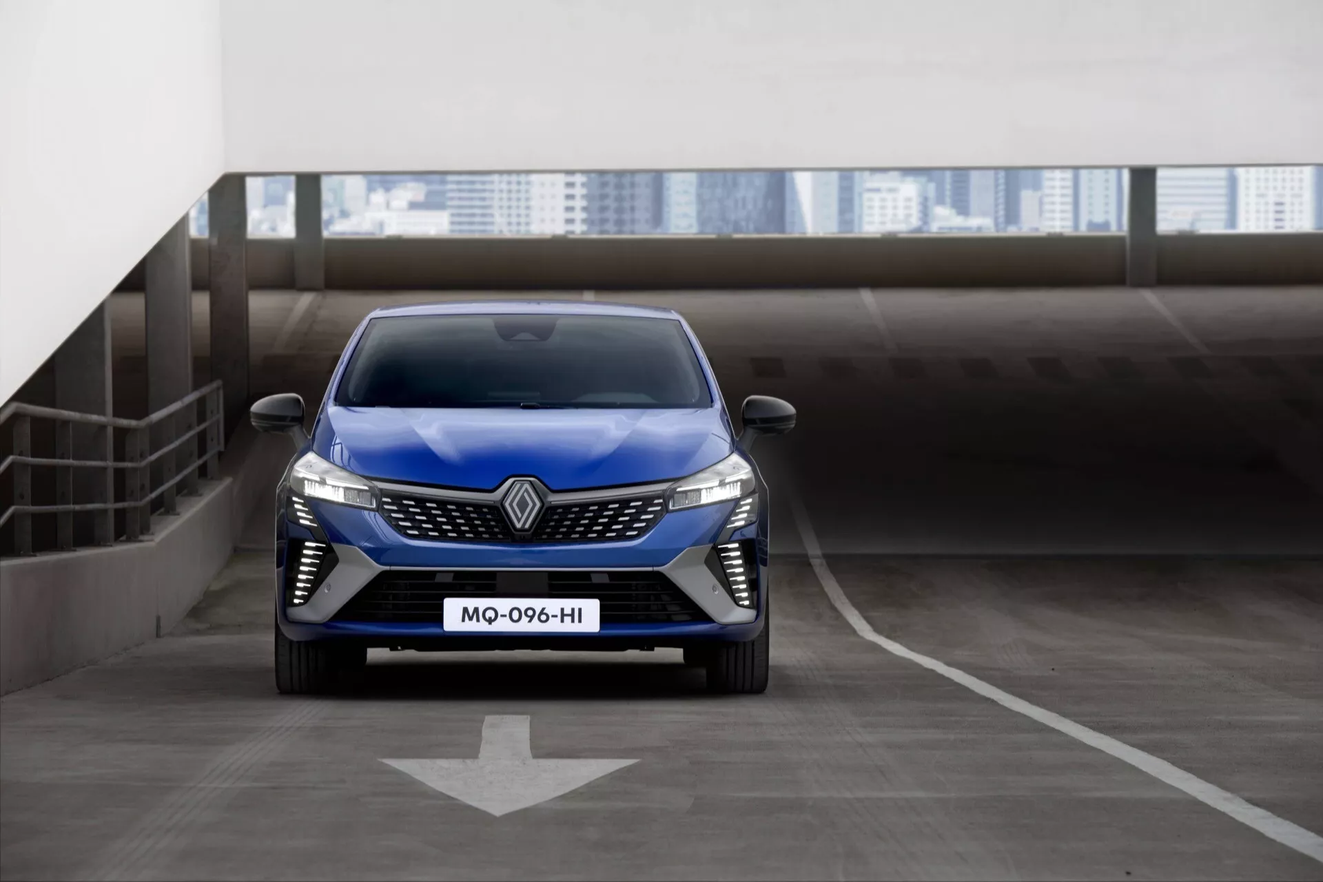Renault Clio V-facelift 2023 en bleu, vue de face sur le sol d'un parking à étages avec flèche directionnelle, présentant des phares LED, une grille proéminente avec un grand logo, et un design de pare-chocs avant sportif