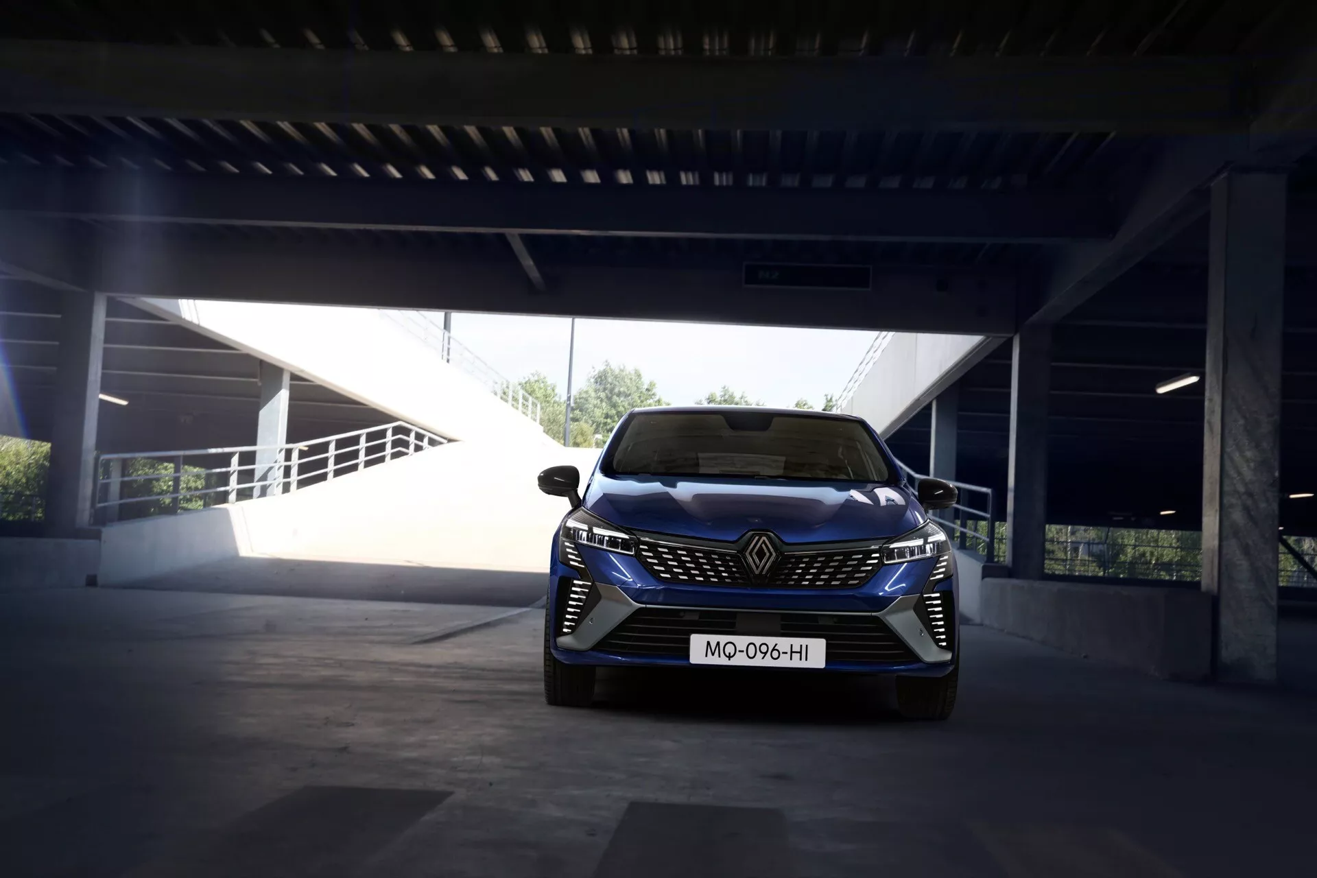 Renault Clio V-Facelift 2023 au jour sur une structure de parking; vue de l'avant montrant l'emblème de la marque, les phares LED, le pare-chocs sportif avec prise d'air et les capteurs d'aide à la conduite.