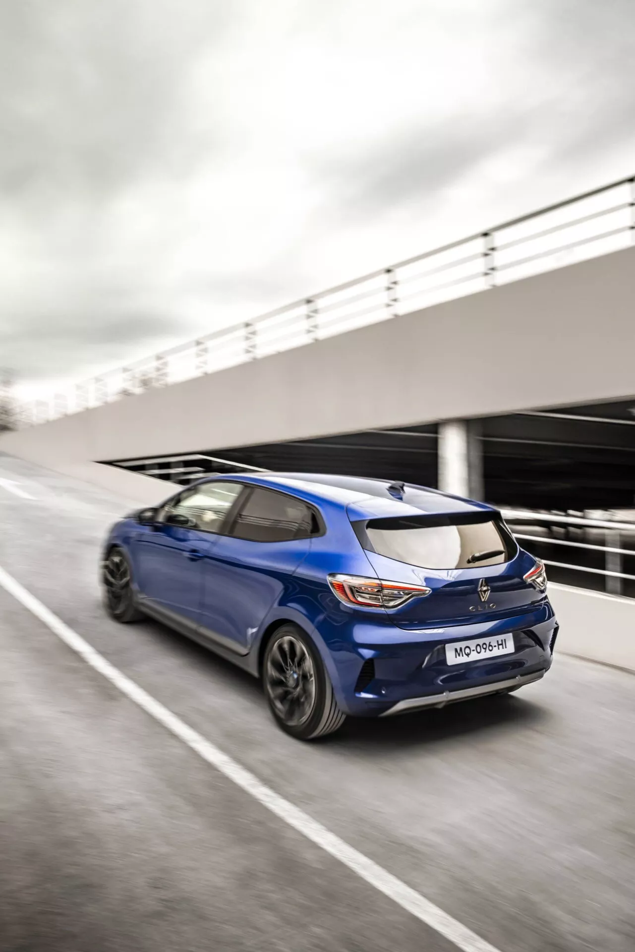 Vue arrière dynamique trois quarts de la Renault Clio V-Facelift 2023 en mouvement sur route, montrant design sportif, feux arrière LED, spoiler monté sur le toit et jantes à multiples branches