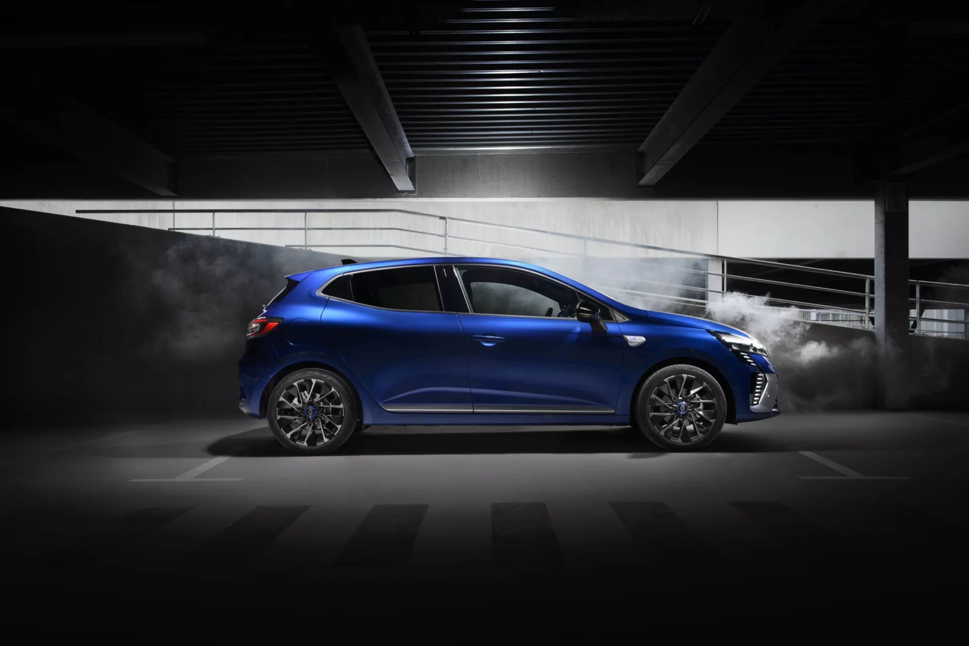 Renault Clio V-Facelift 2023 en bleu brillant avec profil latéral dynamique, prise de vue en contre-plongée mettant en valeur une allure sportive, des lignes de design aérodynamique, des roues en alliage à plusieurs branches et des effets suggérant la vitesse, dans un cadre intérieur dramatiquement éclairé