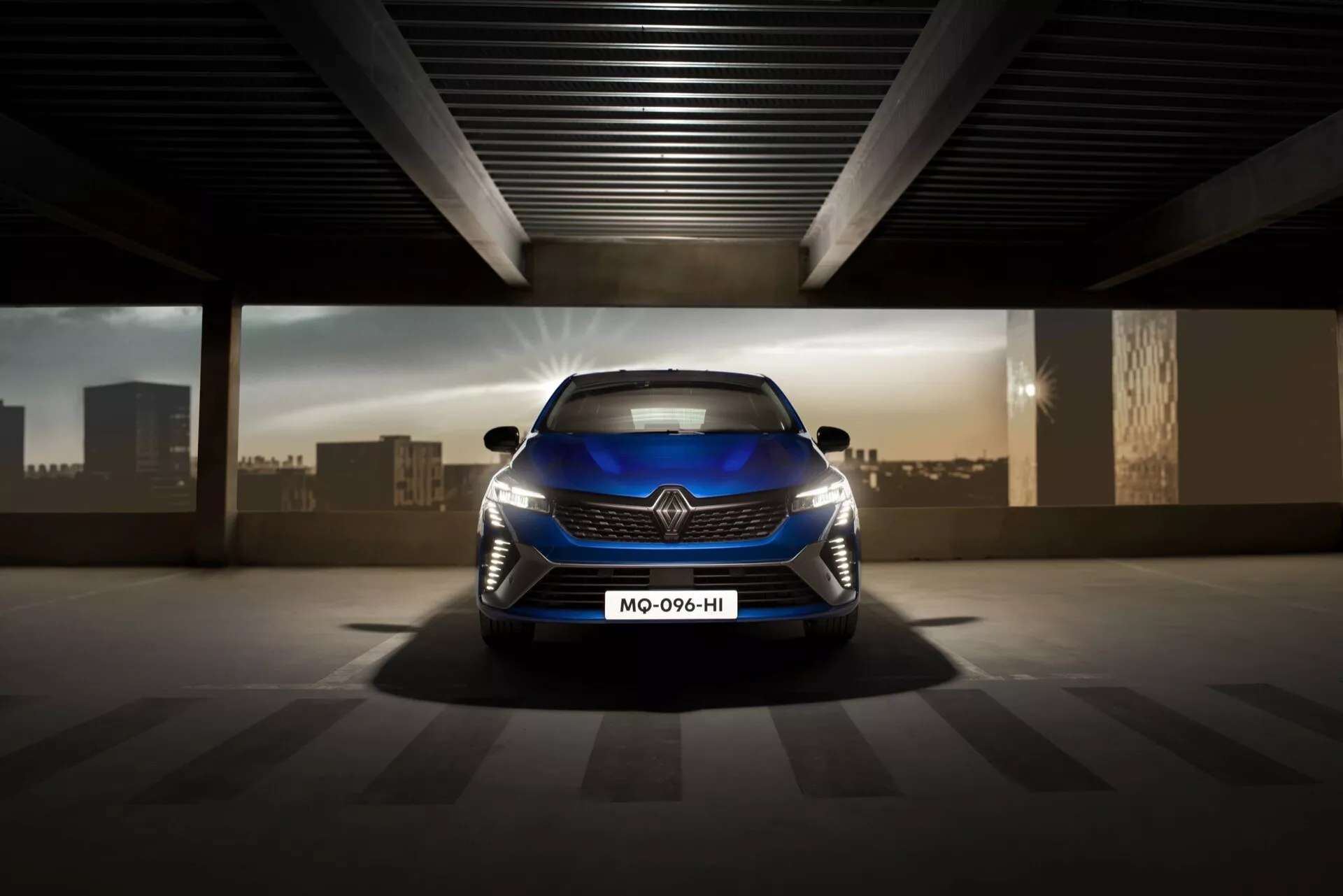 Image de vue avant de la Renault Clio V-facelift 2023 mettant en lumière sa grille sportive, ses phares LED et son design aérodynamique dans une zone de parking éclairée artificiellement