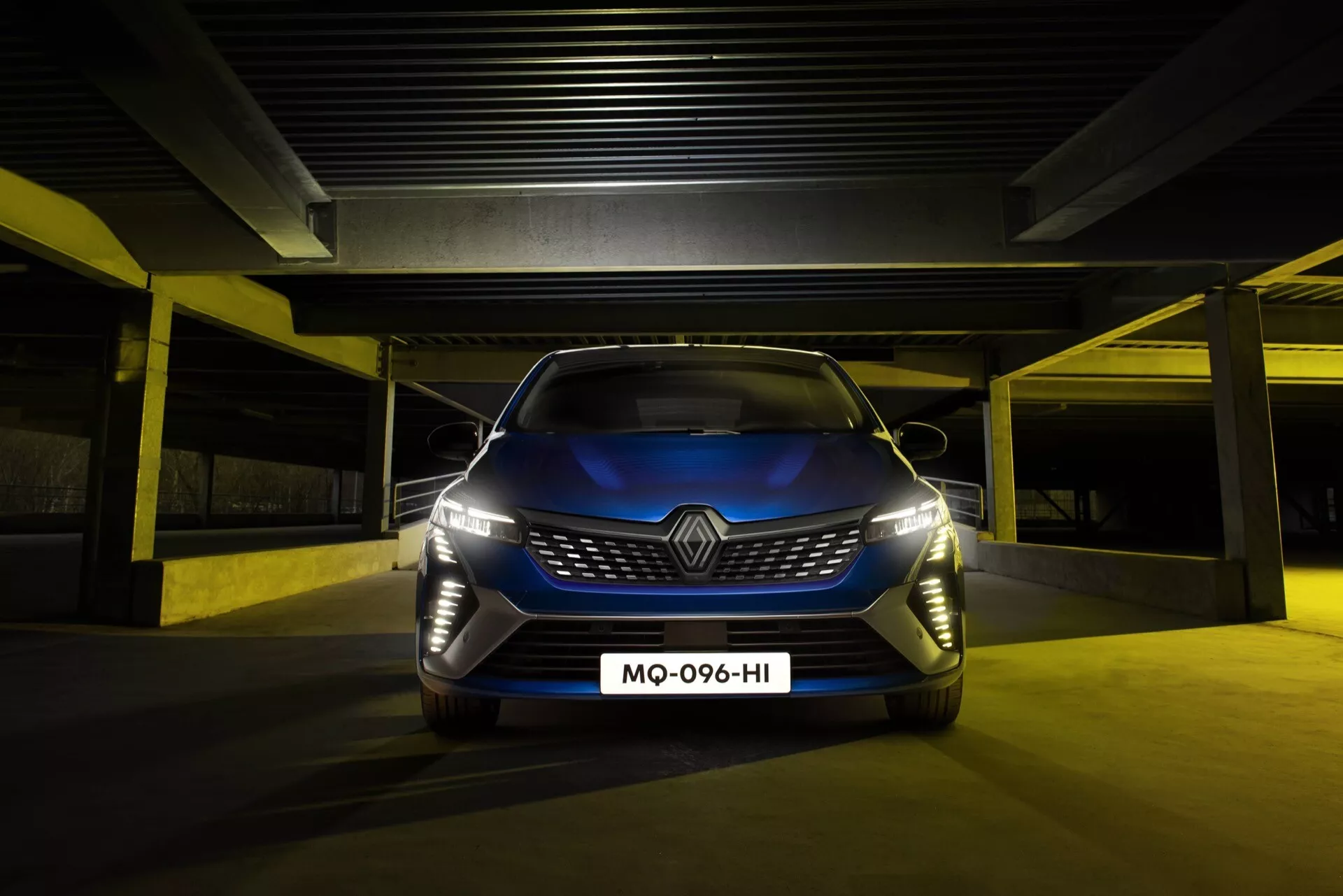 Renault Clio V-Facelift 2023 affichant sa grille audacieuse et ses phares LED sophistiqués depuis un angle avant bas, mettant en lumière les caractéristiques avancées de design et de technologie, dans un cadre de parking faiblement éclairé.