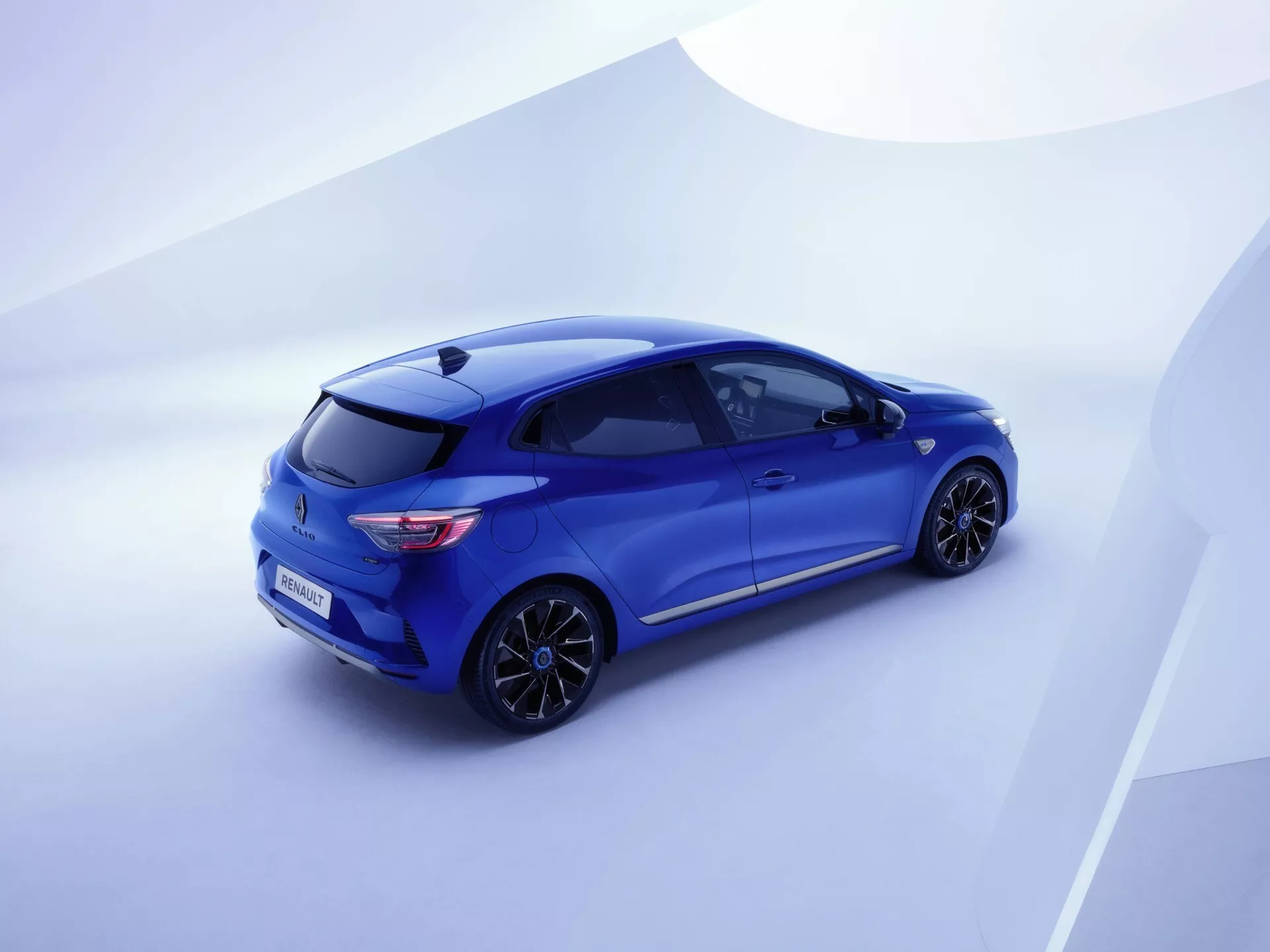Vue arrière en plongée trois-quarts de la Renault Clio V-facelift 2023 en bleu, mettant en évidence ses feux arrière LED, capteurs de stationnement arrière, jante en alliage sportif et caméra de recul intégrée sur un fond dégradé doux