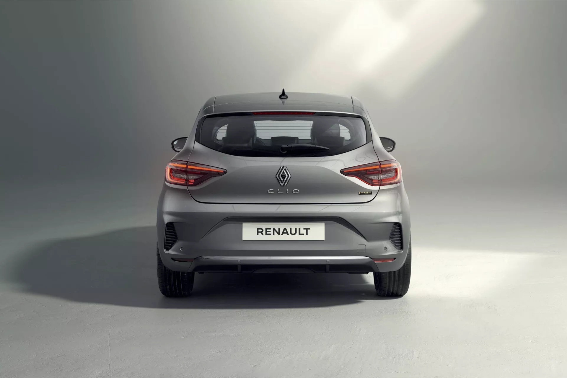 Vue arrière de la Renault Clio hatchback 2023 présentant des feux arrière à LED en forme de C, logo Renault central et badging Clio sur le hayon, pare-brise arrière avec dégivreur, réflecteurs intégrés au pare-chocs arrière et capteurs de stationnement, conception de système d'échappement dissimulé, vue partielle sur les roues en alliage, finition grise métallique avec effet d'ombre dégradée, et une antenne en forme d'aileron de requin, illustrant les avancées en matière de design automobile, de technologie d'éclairage LED et de fonctionnalités d'assistance au conducteur.