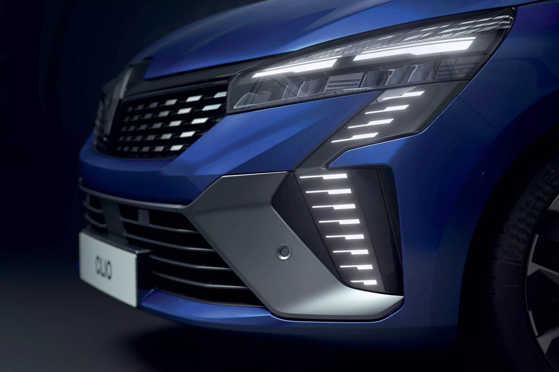 Vue rapprochée de l'avant de la Renault Clio v-facelift 2023, mettant en évidence la grille du côté gauche, phare LED moderne avec feux de jour nets, prise d'air et finition métallique bleu profond