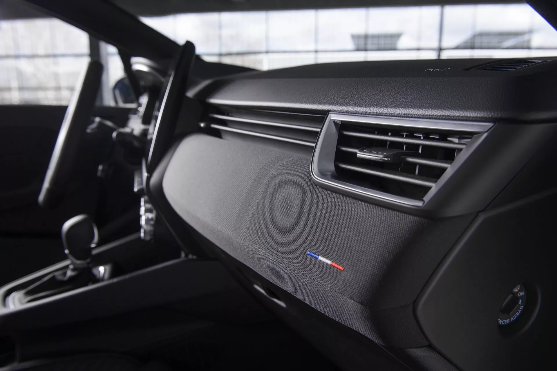 Vue intérieure du Renault Clio V-facelift 2023 depuis le siège du passager, mettant en avant le tableau de bord épuré au design texturé, la ventilation aérodynamique, l'indicateur de contrôle climatique bi-zone, une partie du cadre de la porte et des détails de la console centrale