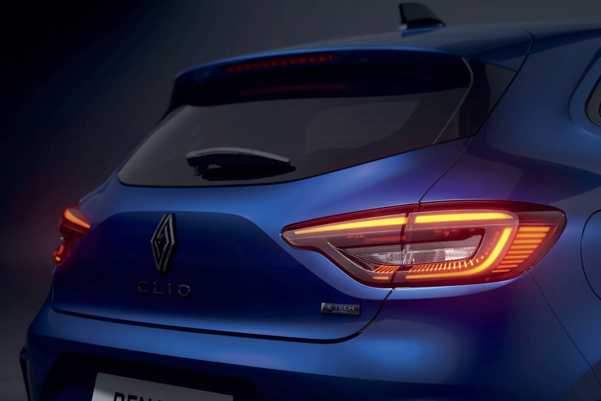 Vue rapprochée d'une Renault Clio V-facelift 2023 montrant le feu arrière LED en forme de C illuminé, détail du badge, et design arrière élégant en bleu métallique sous un éclairage de studio