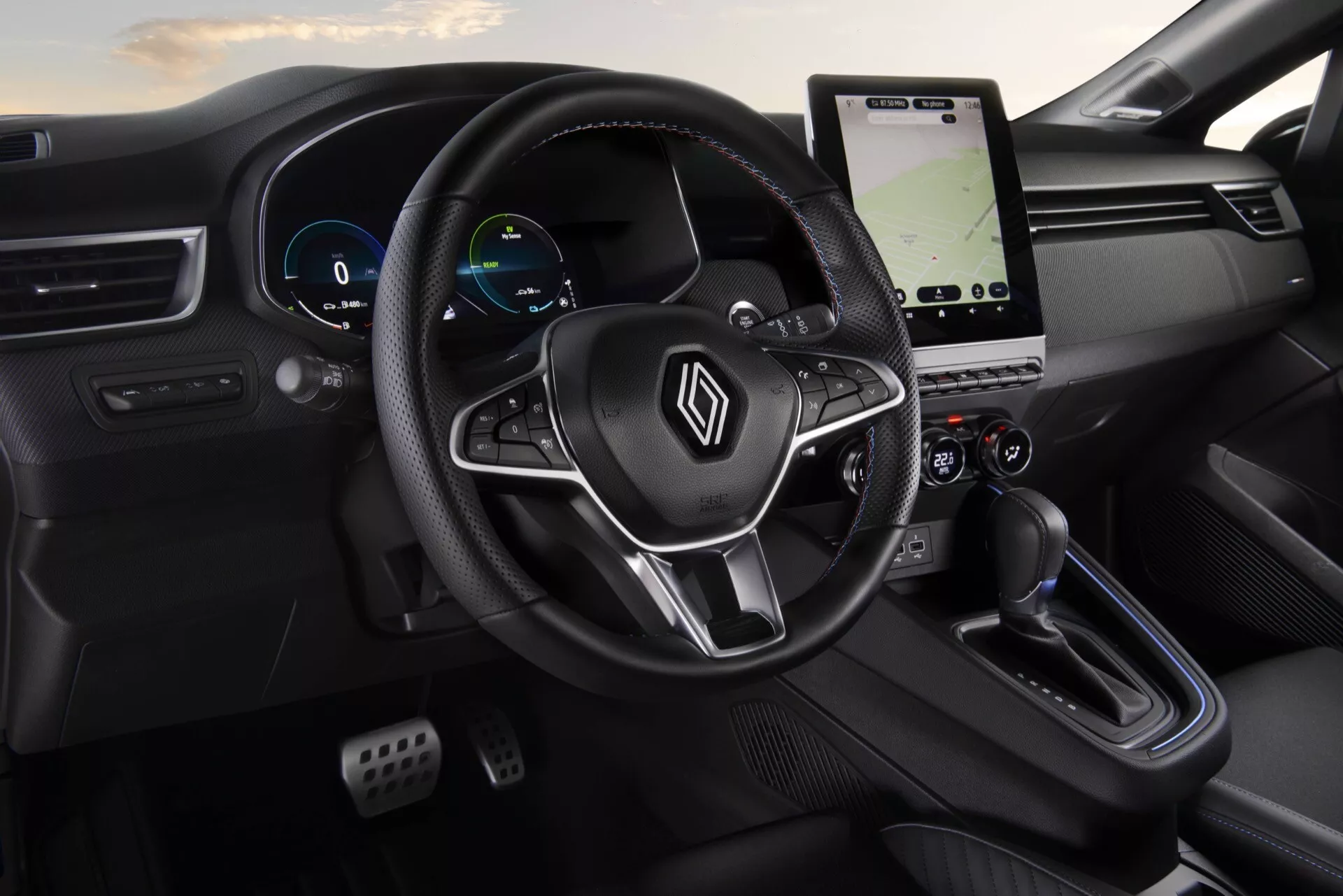 Vue intérieure du Renault Clio V-Facelift 2023 présentant un volant avec logo et boutons multifonctions, tableau de bord numérique, système d'infodivertissement tactile vertical avec navigation, panneau de commande de climatisation, sélecteur de vitesse moderne et design minimaliste élégant en noir du point de vue du passager