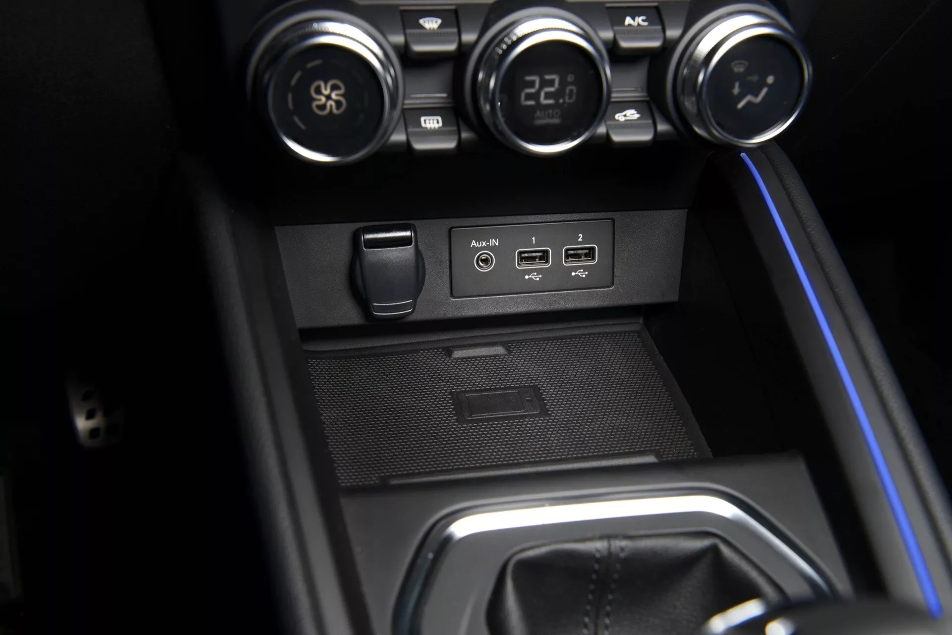 Gros plan sur la console centrale de la Renault Clio V-Facelift 2023 avec boutons de contrôle climatique, ports USB et AUX, prise de courant 12V, compartiment de rangement encastré et zone de levier de vitesse avec éclairage d'ambiance bleu