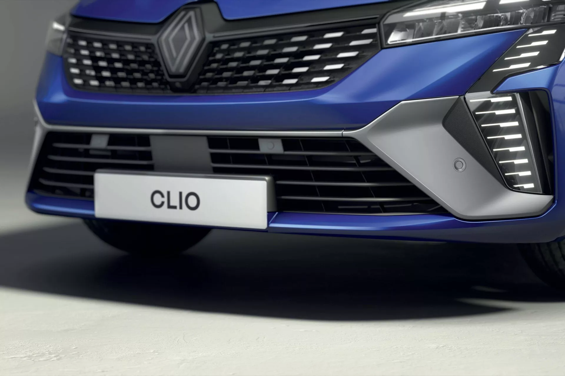 Vue rapprochée, sous un angle bas, de la section avant de la Renault Clio V-facelift 2023, se concentrant sur la grille, les entrées d'air et la conception du pare-chocs, avec une finition métallique bleue brillante et un éclairage avancé