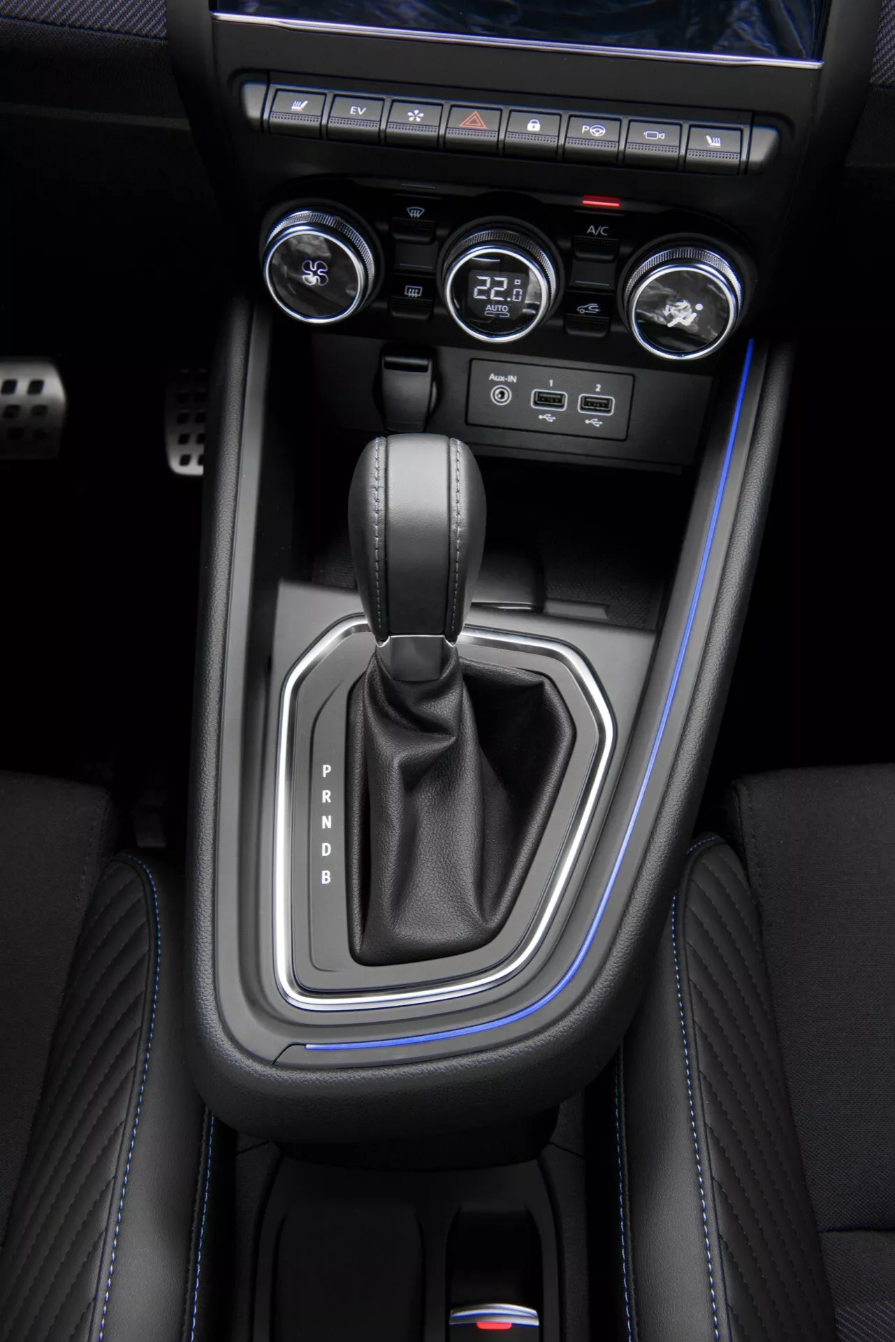 Vue aérienne de la console centrale du Renault Clio V-Facelift 2023 montrant le levier de vitesse en position Park avec panneau argenté, coutures en cuir noir, boutons de contrôle climatique et système de démarrage sans clé