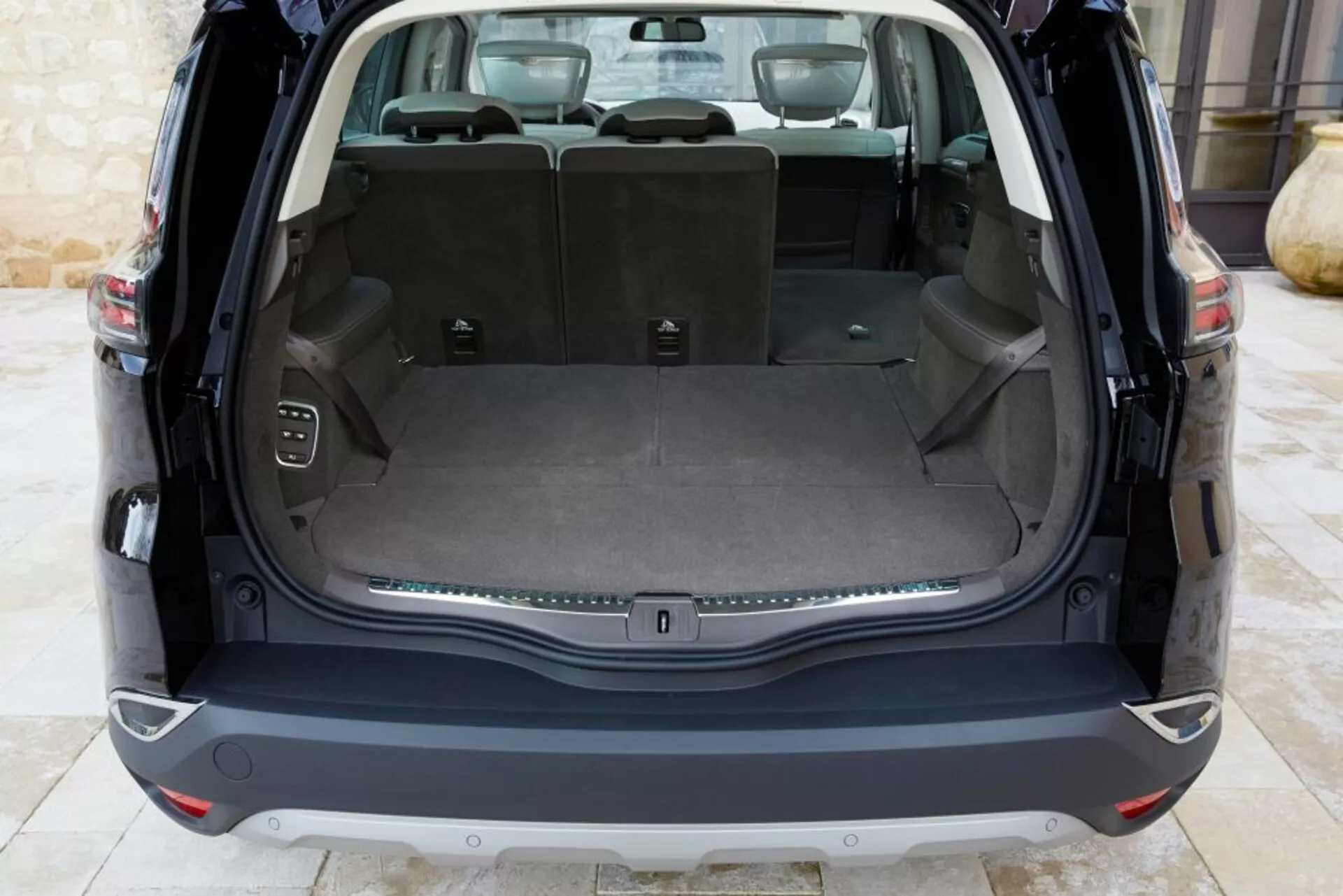 Vue intérieure arrière d'une Renault Espace 2015 montrant un espace de chargement propre avec le hayon ouvert, comportant un plancher de chargement plat, des sièges arrière rabattables 60/40, une prise de courant, des crochets pour le chargement, un bouton de commande pour le hayon électronique, et une bande décorative chromée, sous un éclairage naturel.