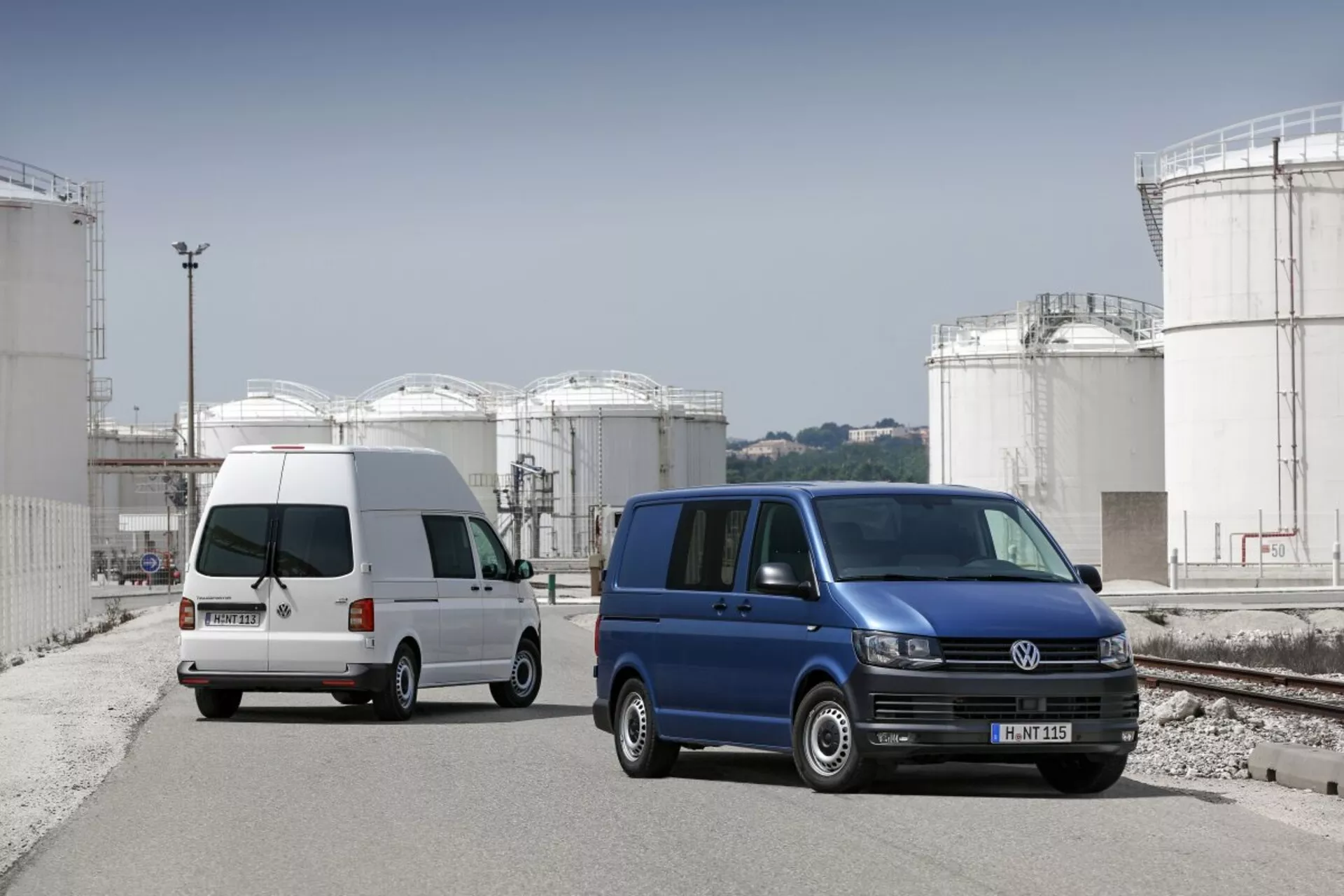 Deux fourgonnettes Volkswagen Transporter garées sur un site industriel avec de grands réservoirs de stockage en arrière-plan, montrant leur conception utilitaire commerciale, portes latérales coulissantes et roues en alliage standard.