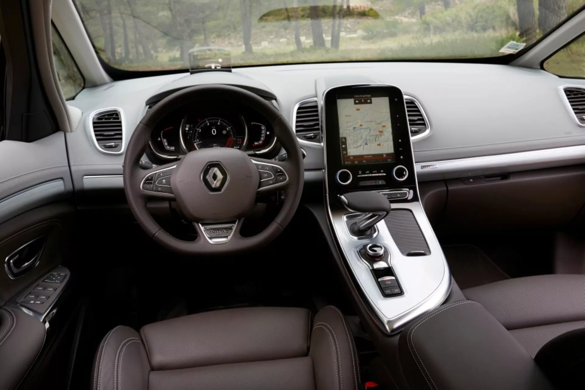 Vue intérieure du Renault Espace 2015 depuis la place du conducteur, présentant un design luxueux avec un volant à trois branches avec boutons de commande, un tableau de bord numérique, un écran tactile vertical sur la console centrale pour le système d'infodivertissement, un levier de vitesse automatique, des sièges en cuir avec réglage électronique, et des aérations sur le tableau de bord pour la climatisation