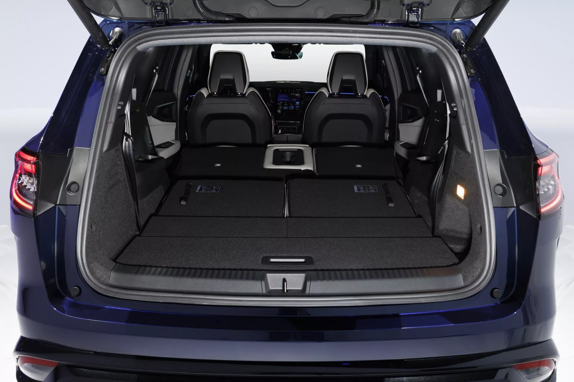 Vue de l'espace de chargement du Renault Espace VI 2023 avec porte arrière ouverte montrant un coffre spacieux, un plancher de chargement plat, des points d'attache pour couvercle de cargaison, des compartiments de rangement latéraux, un intérieur moquetté, des porte-gobelets et des lumières d'illumination