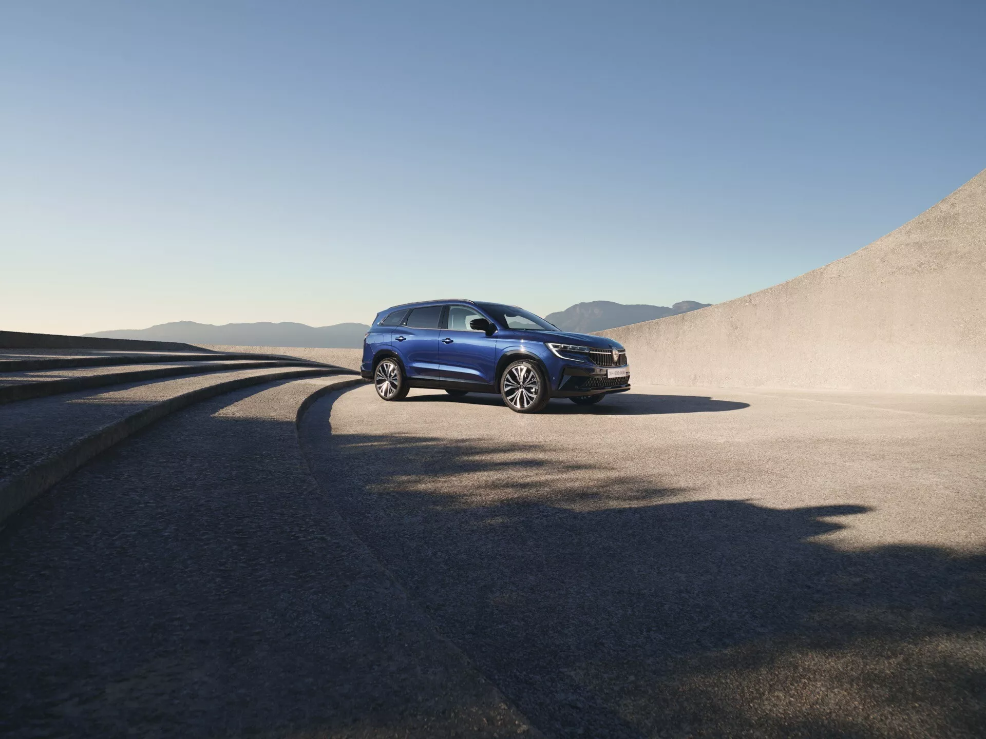 SUV Renault Espace VI 2023 en finition métallique sur route ouverte avec arrière-plan montagneux, mettant en évidence la garde au sol et le design aérodynamique, doté de phares LED, de jantes en alliage à multiples branches et de l'emblème de la marque sur la calandre