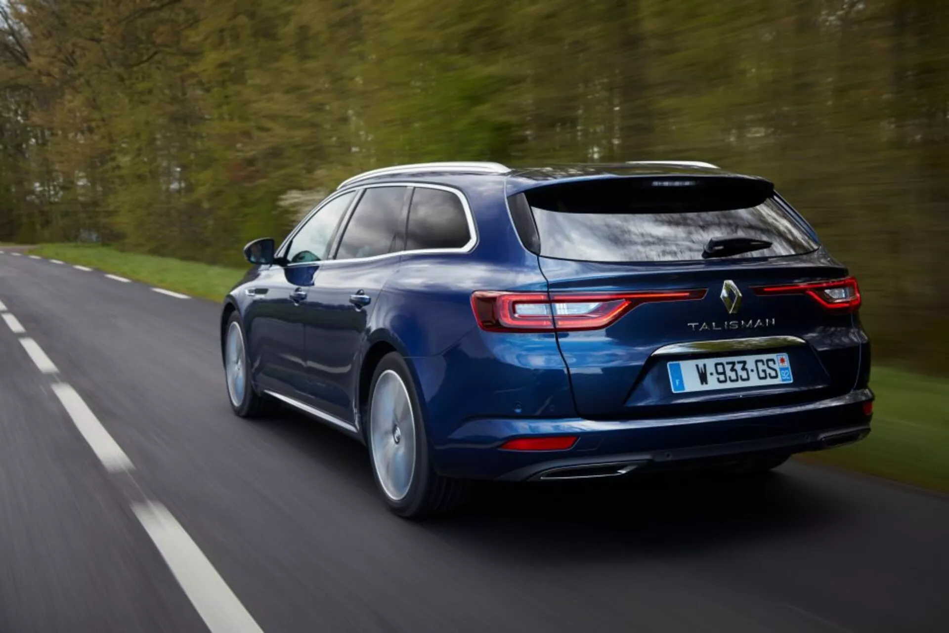Break bleu Renault Talisman Grandtour 2016 en mouvement, présentant des caractéristiques de conception telles que des feux arrière LED, des tuyaux d'échappement et sa silhouette élégante depuis un angle arrière et légèrement surélevé sur une route bordée d'arbres.