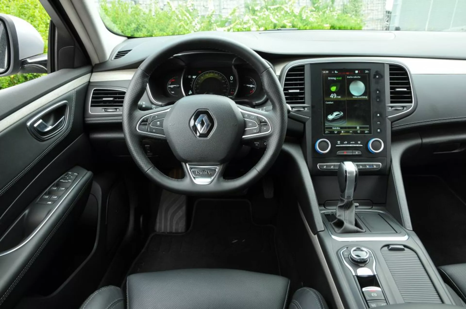 Intérieur du Renault Talisman 2016 montrant le cockpit du conducteur avec un volant multifonction en cuir, tableau de bord numérique, système d'infodivertissement tactile vertical et commandes de climatisation élégantes depuis la perspective du passager arrière